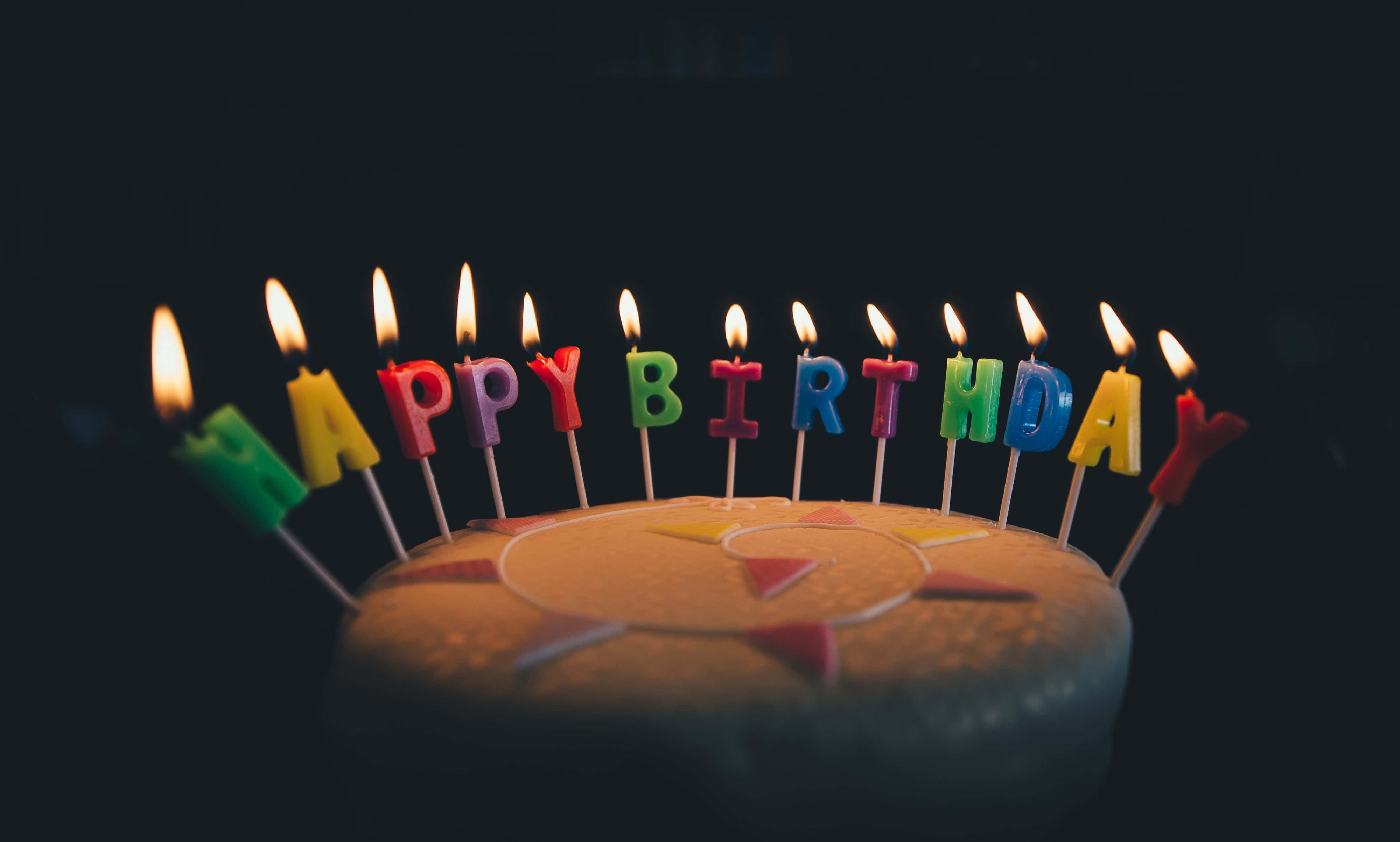 Cute Birthday 桌布 壁紙 — 一個生日蛋糕，上面有彩色字母蠟燭拼出「生日快樂」，在黑暗背景中閃耀。