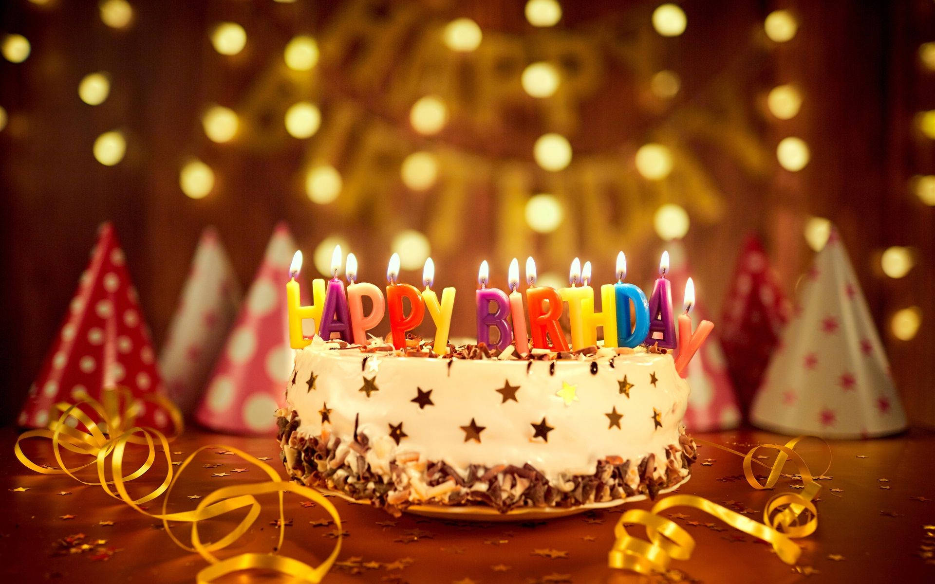 生日快樂 桌布 壁紙 — 一個生日蛋糕，上面有彩色蠟燭拼出 happy birthday，周圍有派對帽和彩帶，在溫暖的燈光下。