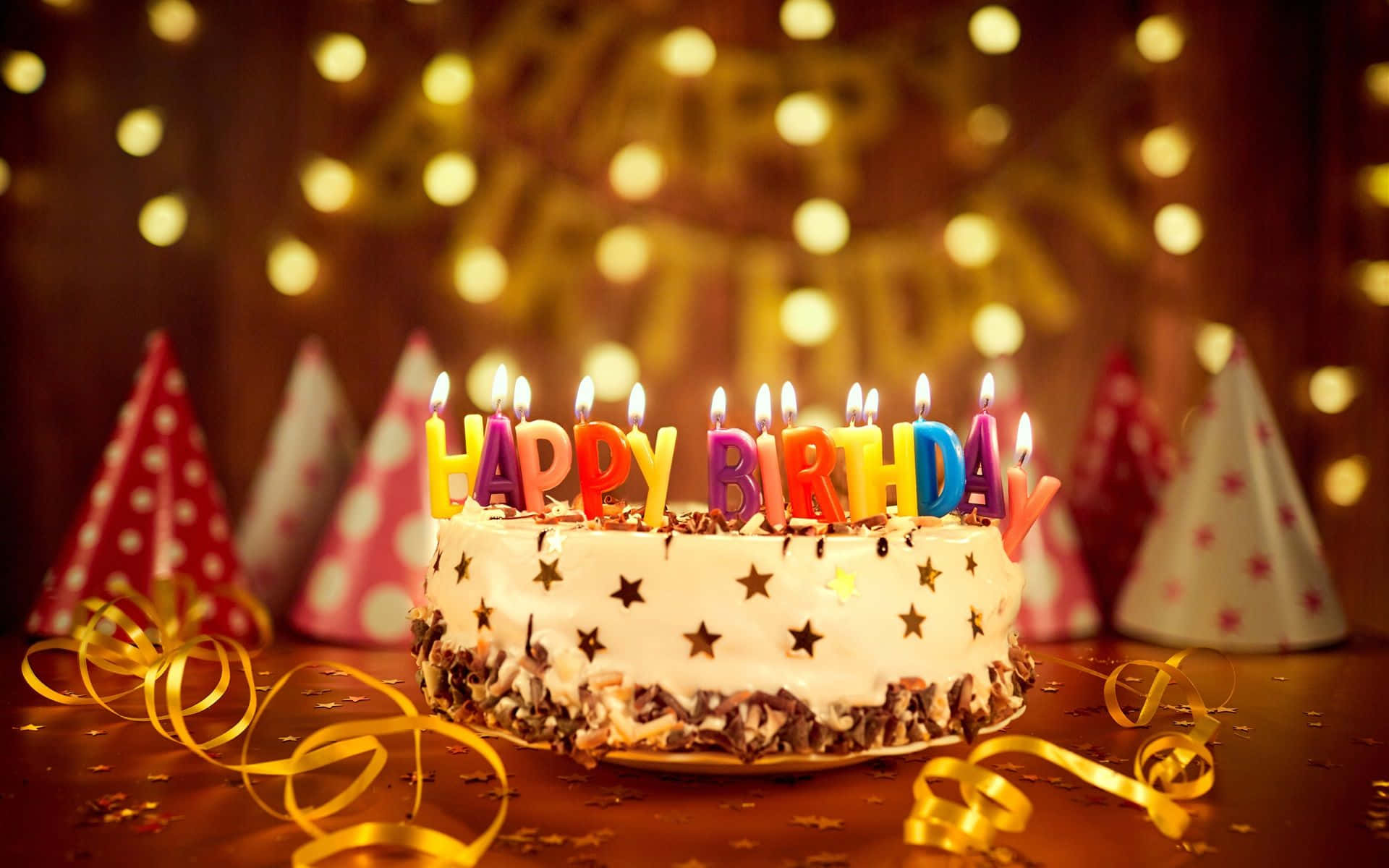 Birthday 桌布 壁紙 — 一個生日蛋糕，上面有彩色蠟燭拼出 happy birthday，周圍環繞著派對帽和金色彩帶，在溫暖的串燈下。