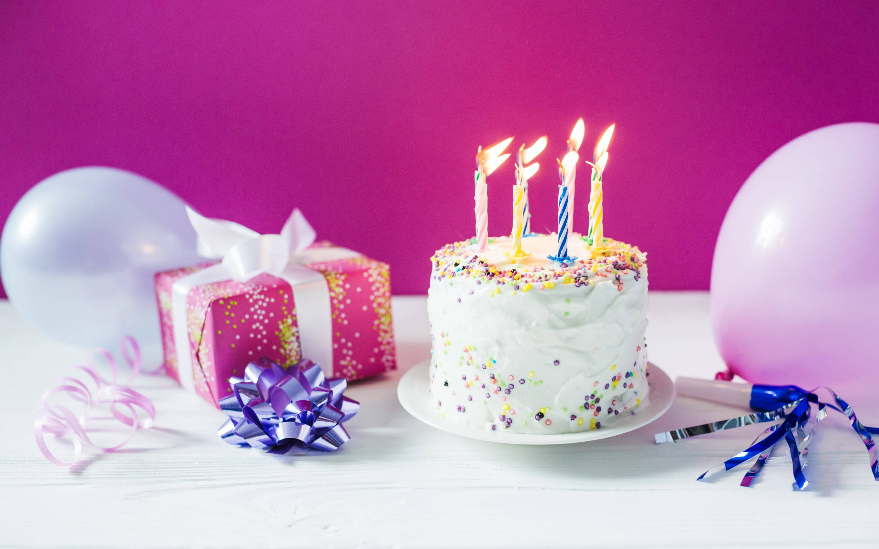 Birthday Cake 桌布 壁紙 — 一個生日蛋糕，上面有燃燒的蠟燭和彩色糖粒，旁邊放著禮物、氣球和彩帶，放在白色桌子上，背景是鮮豔的粉紅色。