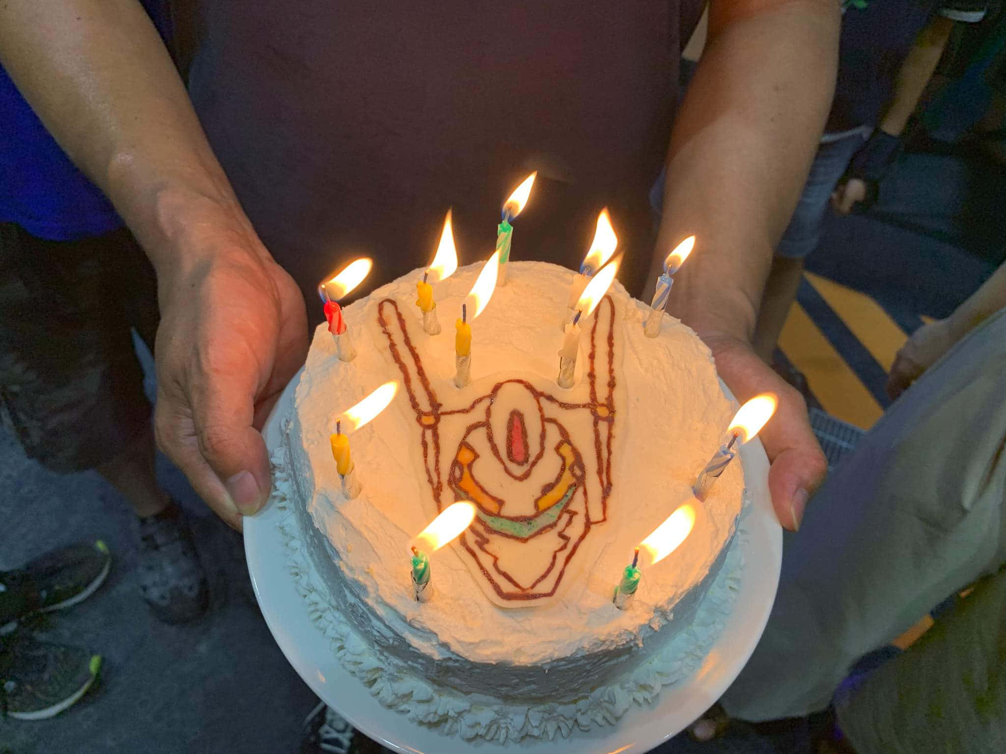 Birthday Cake 桌布 壁紙 — 一個生日蛋糕，上面有燃點著的蠟燭，形狀像機器人臉，由兩隻手在昏暗的房間裡捧著。