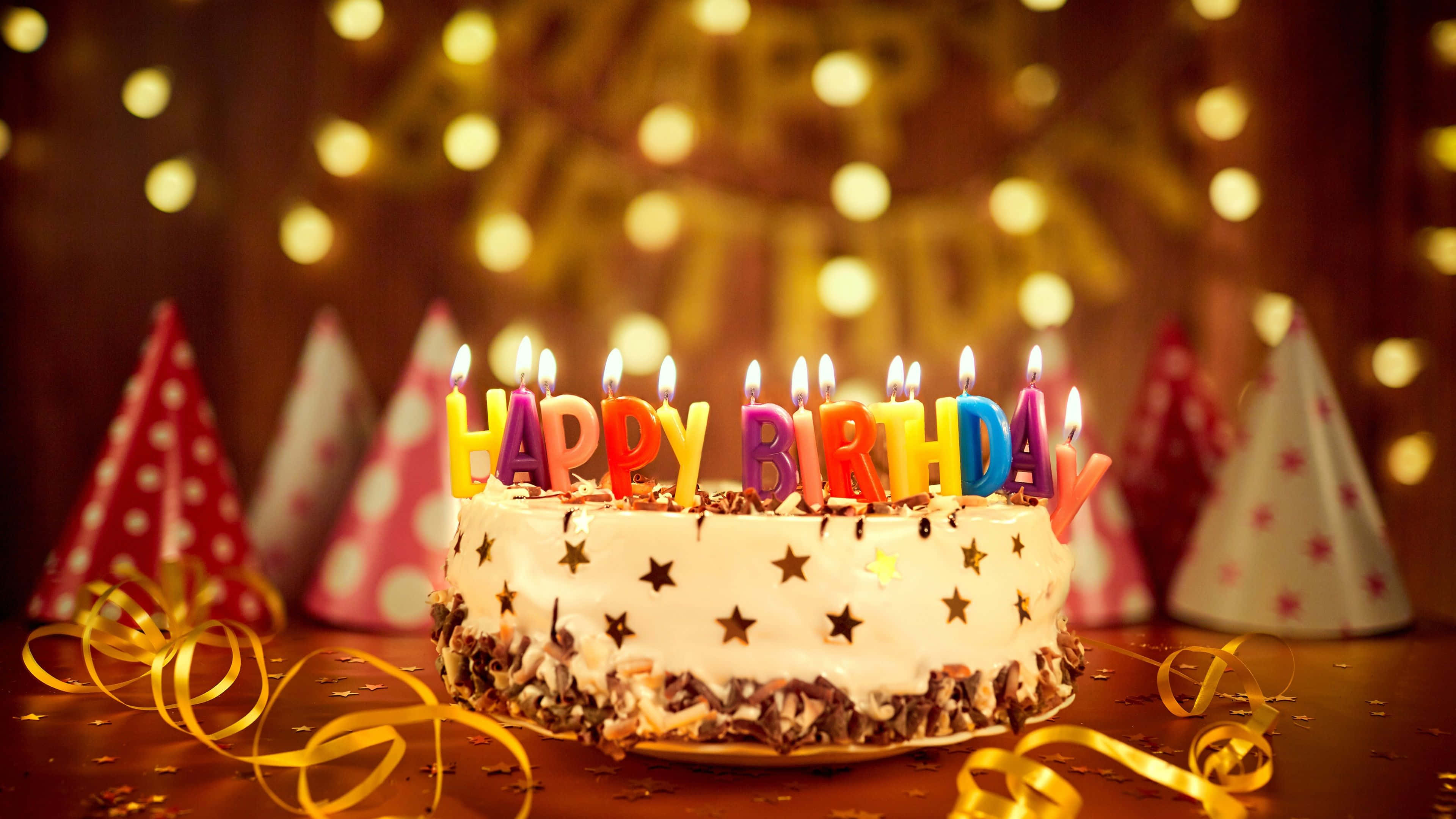 Birthday Cake 桌布 壁紙 — 一個生日蛋糕，上面有色彩繽紛的蠟燭點燃拼出 happy birthday，周圍環繞著派對帽和彩帶，在溫暖的散景燈光下。