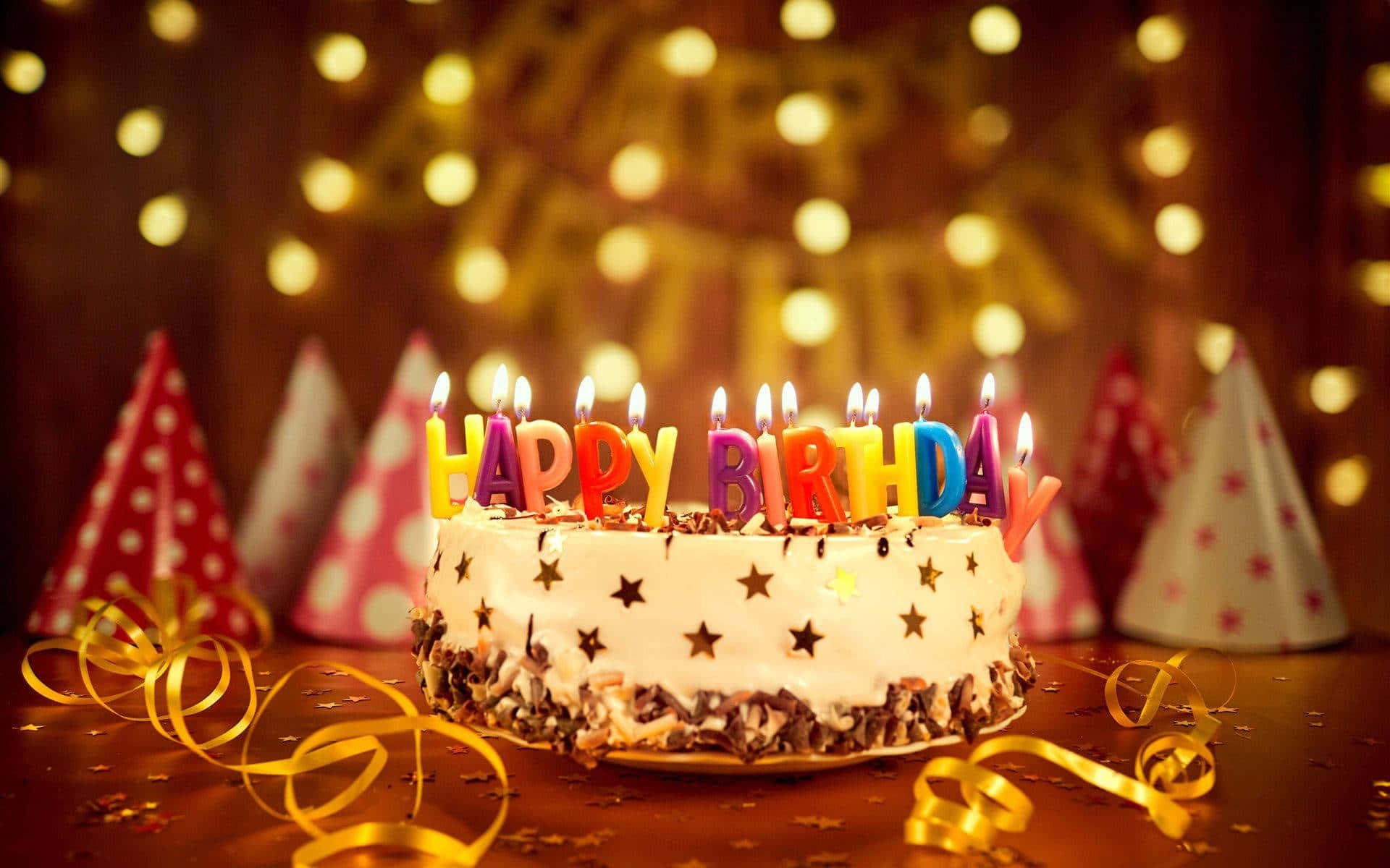 High Resolution Happy Birthday 桌布 壁紙 — 一個生日蛋糕，上面有色彩繽紛的點燃蠟燭拼出 happy birthday，周圍環繞著派對帽和金色彩帶，在溫暖的散景燈光下。