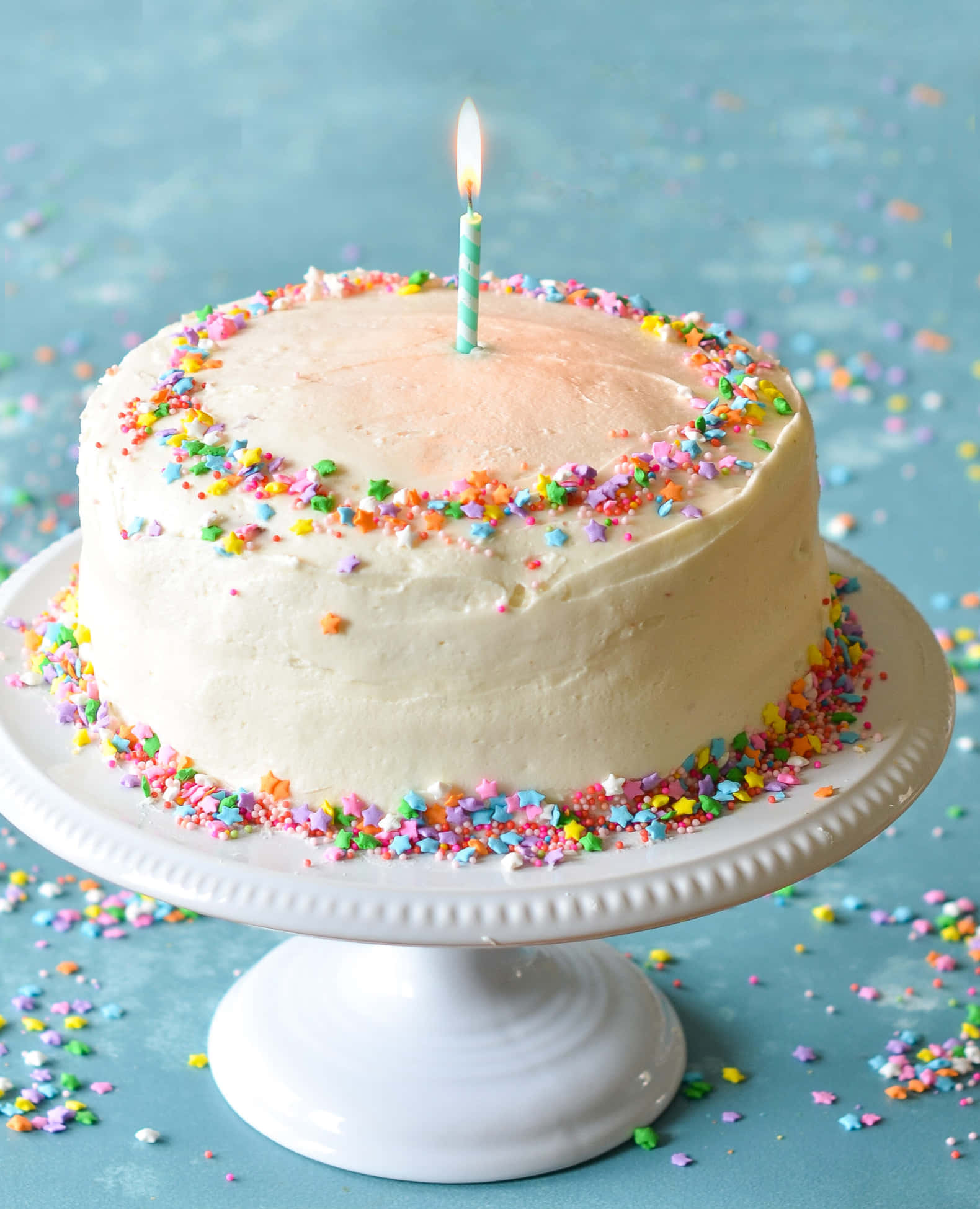 Buttercream 桌布 壁紙 — 一個生日蛋糕，白色糖霜配上彩色星星糖粒，頂部插著點燃的蠟燭，放在白色支架上，背景為藍色。