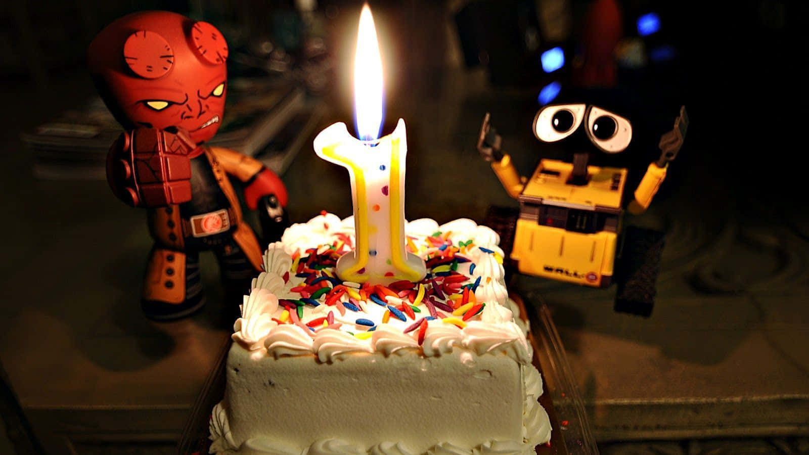搞笑 桌布 壁紙 — 一個生日蛋糕上插著一支點燃的數字一蠟燭，兩側分別是紅色的Hellboy公仔和黃色的Wall-E玩具，處於昏暗的環境中。