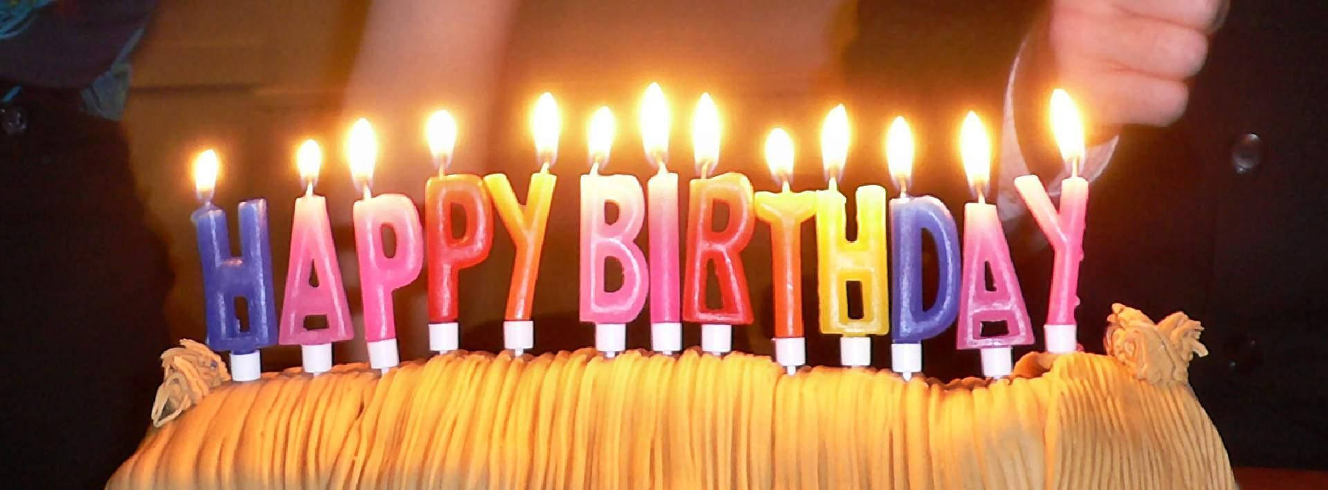 蛋糕 桌布 壁紙 — 一個生日蛋糕的特寫，上面有色彩繽紛的點燃蠟燭，在深色背景下拼出「HAPPY BIRTHDAY」。