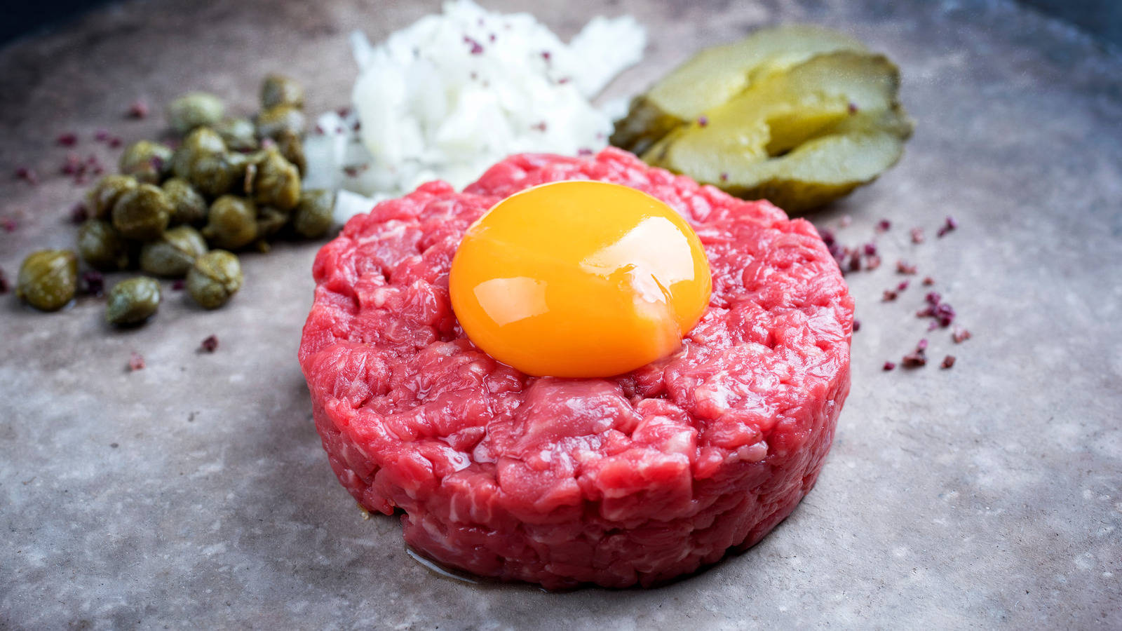 meat 桌布 壁紙 — 一個生牛肉他他肉餅，上面放著一個亮黃色的蛋黃，周圍有酸豆、醃黃瓜和洋蔥，放在有紋理的灰色表面上。