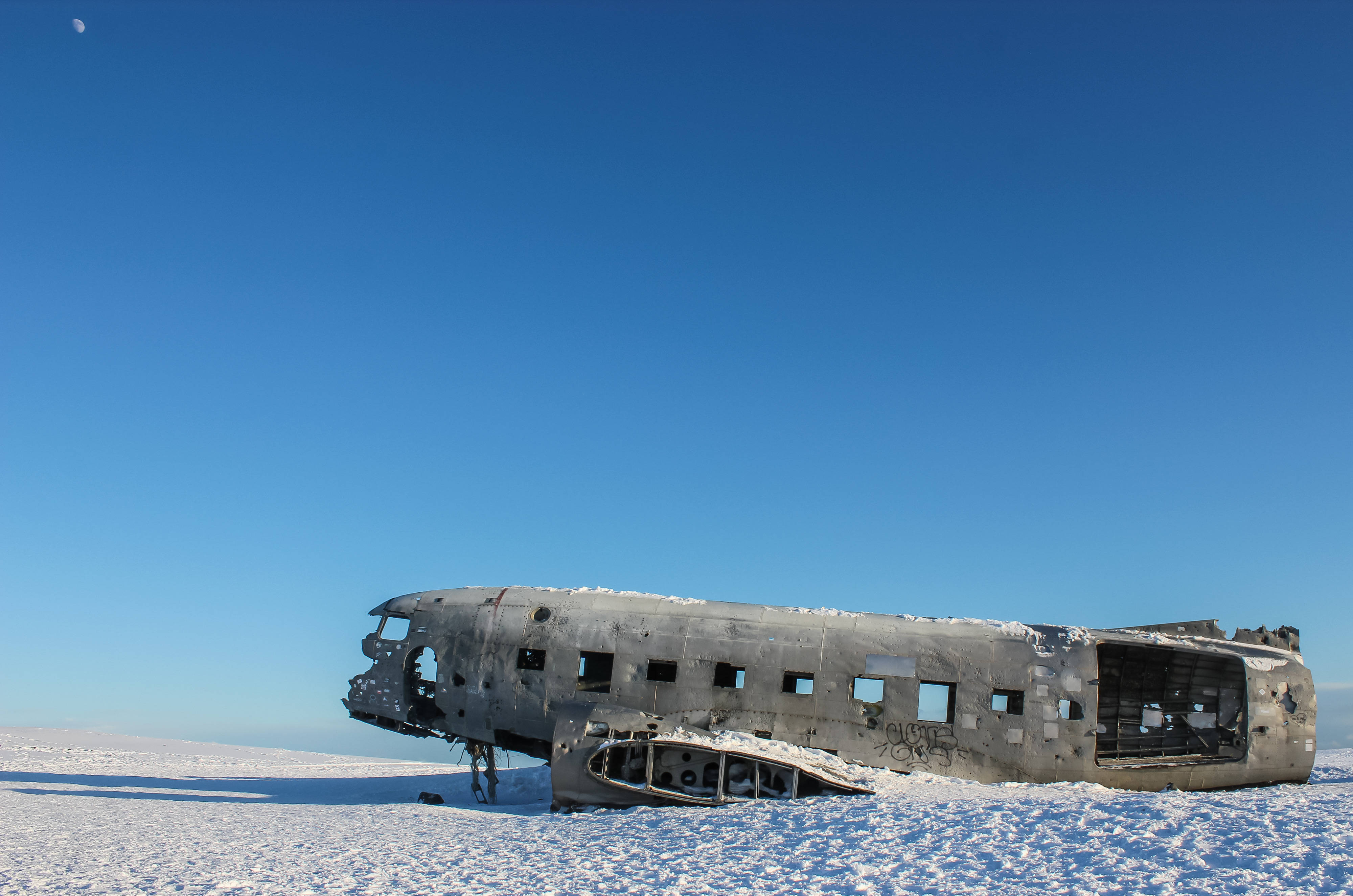 Plane 桌布 壁紙 — 一個生鏽的飛機機身被遺棄在廣闊的雪地上，天空清澈湛藍，可見月亮。