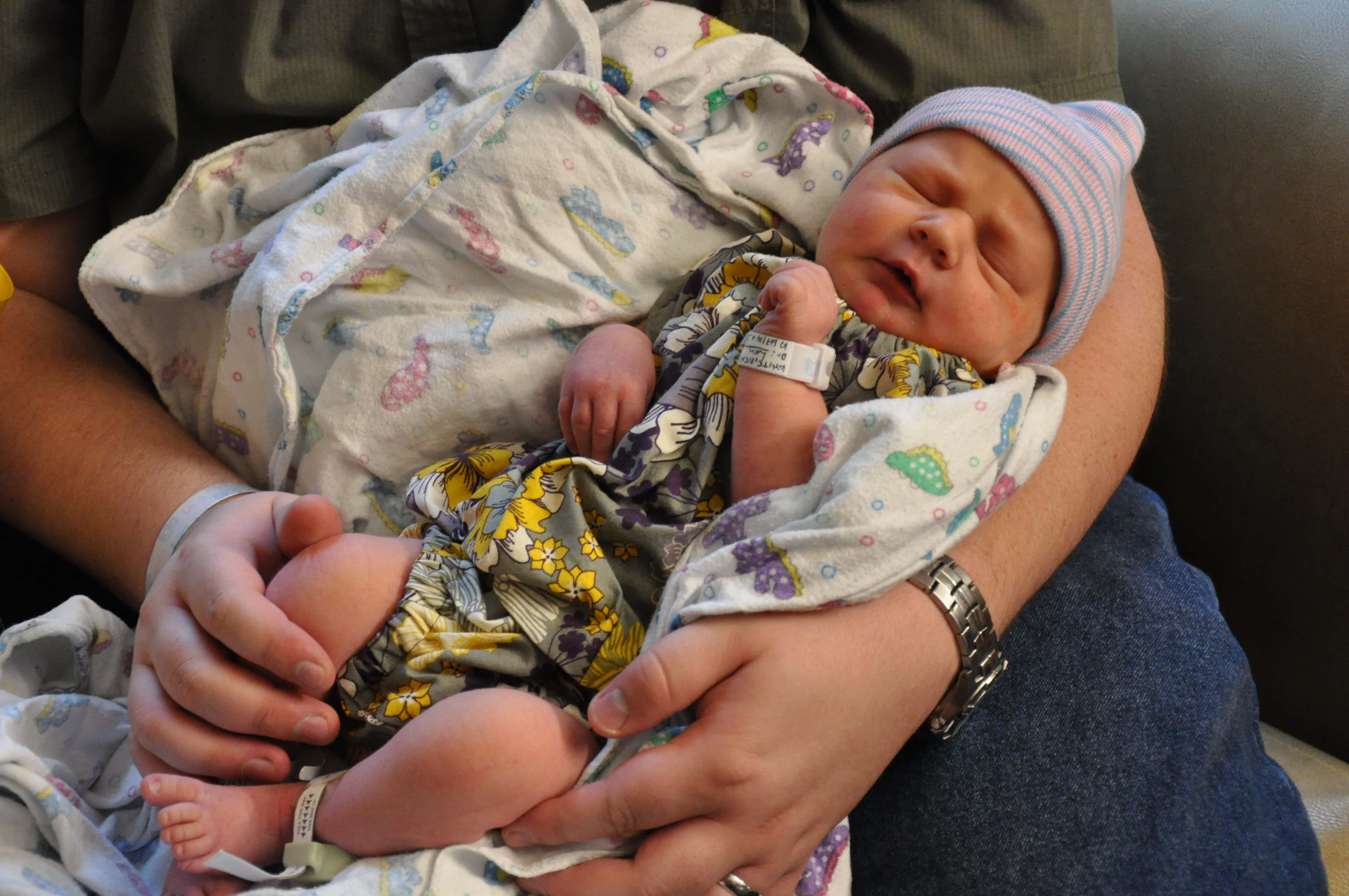 Newborn Baby 桌布 壁紙 — 一個用圖案毛毯包裹的新生嬰兒，戴著條紋帽子和醫院手環，在成人懷中安詳地睡覺。