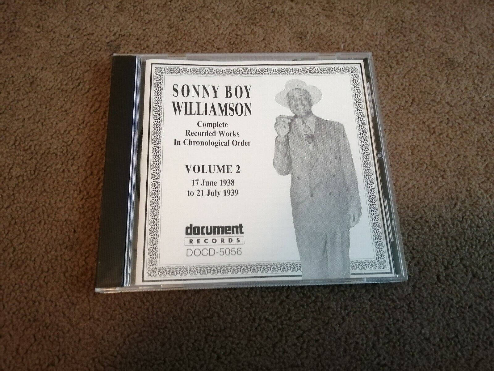 黑白 桌布 壁紙 — 一個用於Sonny Boy Williamson音樂的復古CD盒放在棕色地毯上，上面有藝術家穿著西裝和帽子的黑白照片。