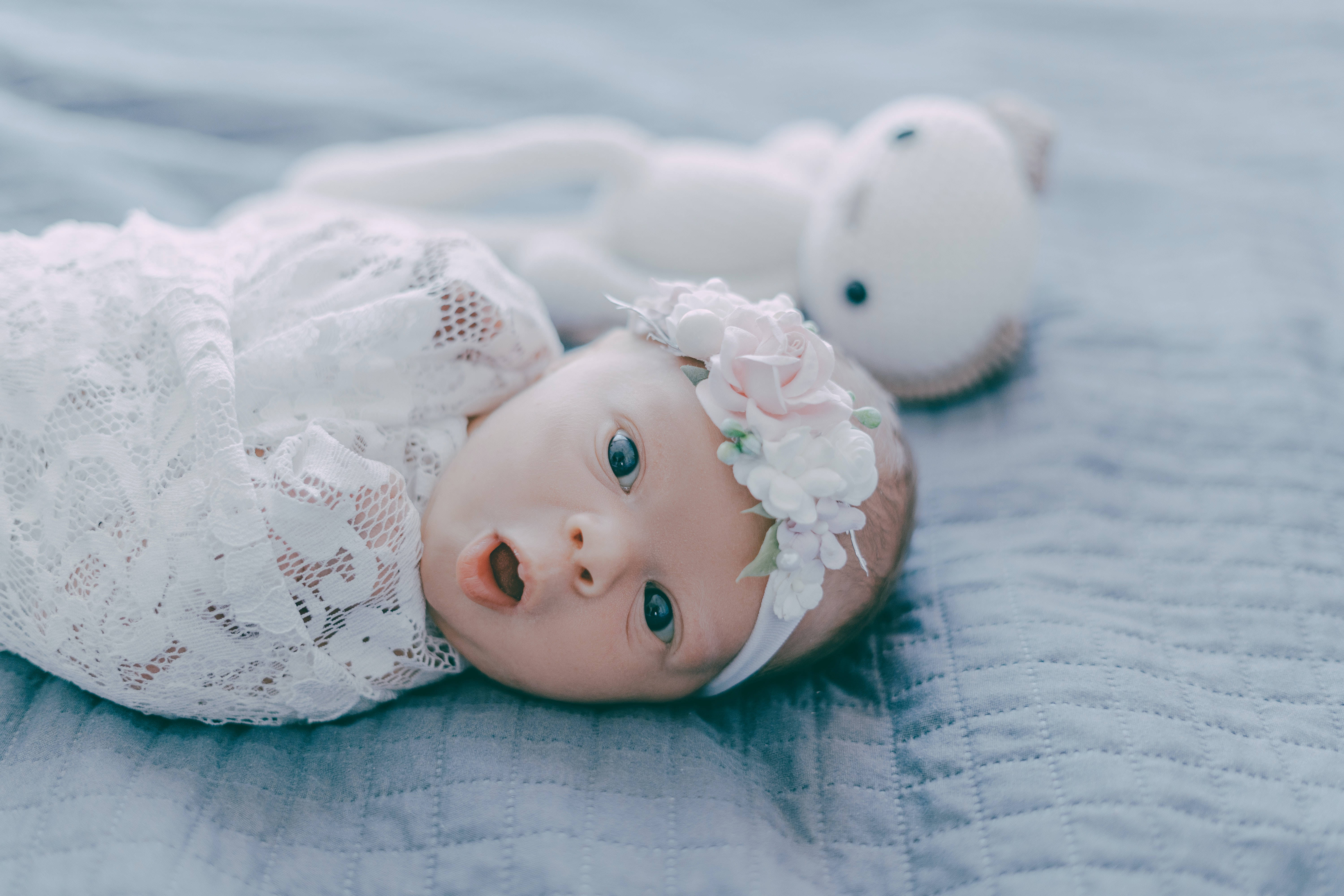 Very Cute Baby 桌布 壁紙 — 一個用白色蕾絲包裹的新生嬰兒躺在柔軟的藍色羽絨被上，旁邊有一隻柔軟的白色泰迪熊。