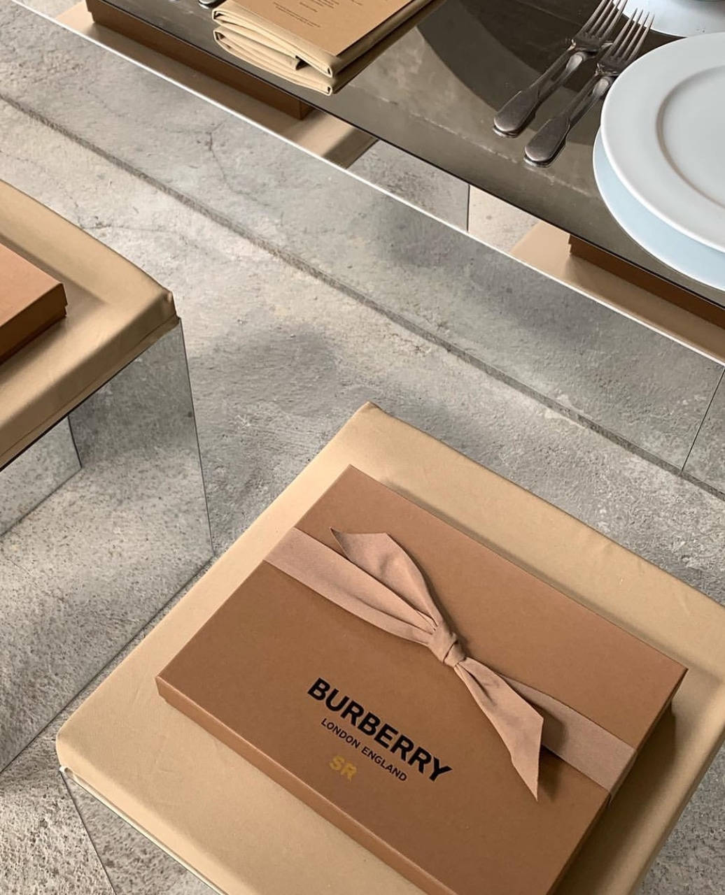 Burberry 桌布 壁紙 — 一個用米色絲帶繫著的 Burberry 禮物盒放在現代混凝土桌上，旁邊有疊起的盤子和餐具。