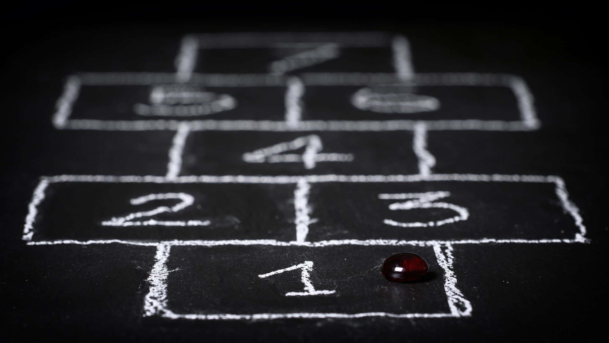 Educational Games 桌布 壁紙 — 一個用粉筆在深色表面上畫的跳房子格子的特寫，一顆紅色彈珠在第一格上。
