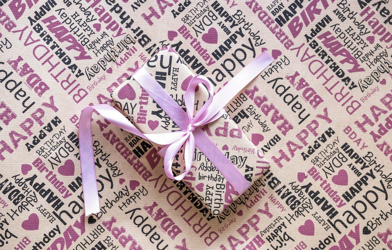 Pink Birthday 桌布 壁紙 — 一個用粉紅色絲帶綁著的禮物盒，放在佈滿重複「happy birthday」文字和心形的生日主題包裝紙上。