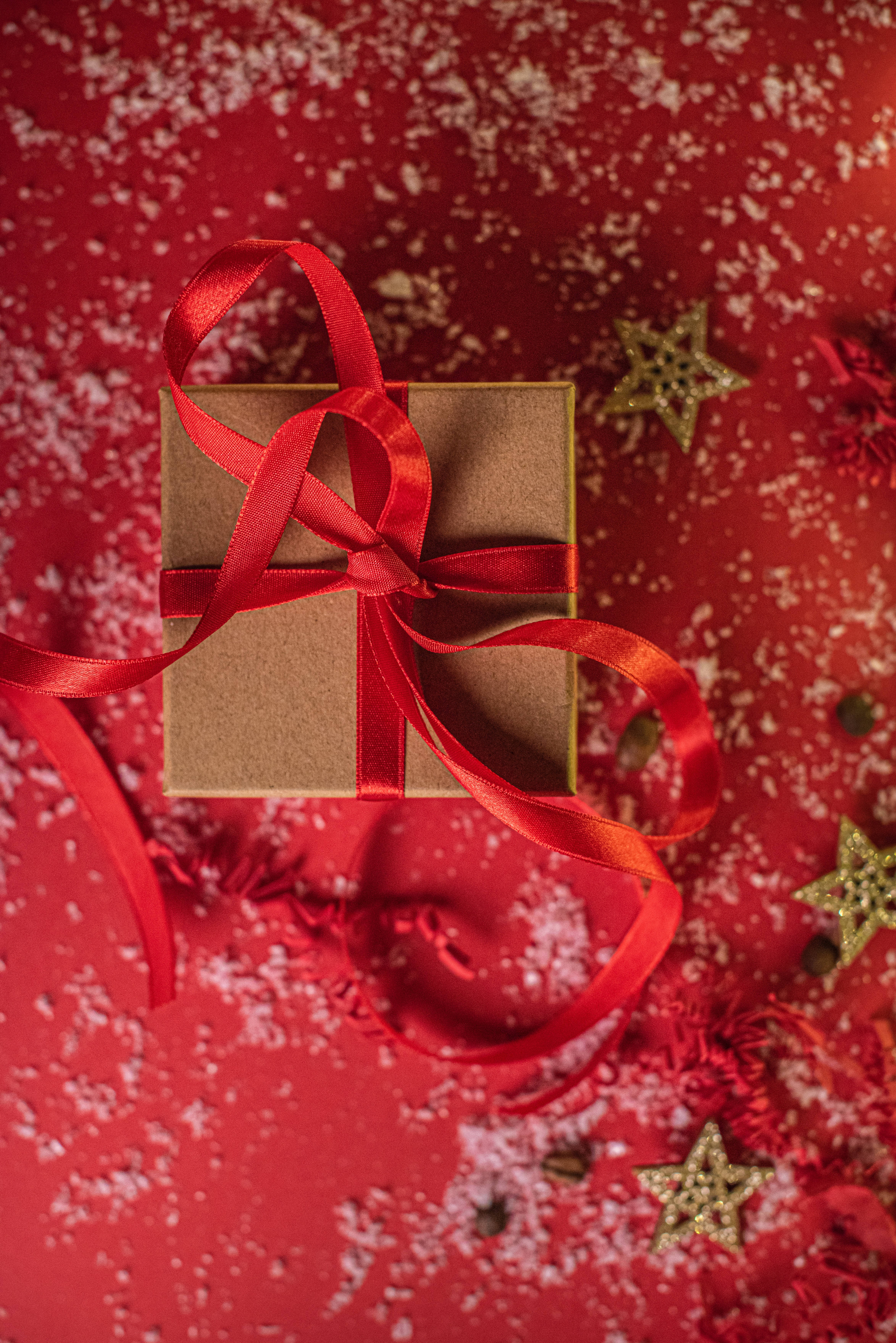 Red Christmas 桌布 壁紙 — 一個用紅色緞帶綁著的棕色禮物盒，放在一個撒滿閃亮星星和假雪的節日紅色表面上。