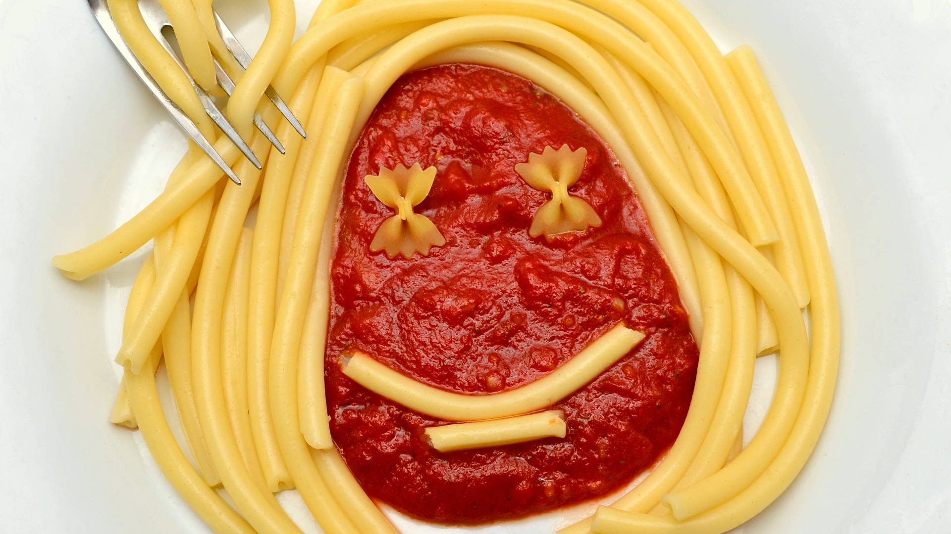 Pasta 桌布 壁紙 — 一個用義大利麵頭髮、蝴蝶結眼睛和麵條微笑在番茄醬中放在白色盤子上的愉快麵食臉。