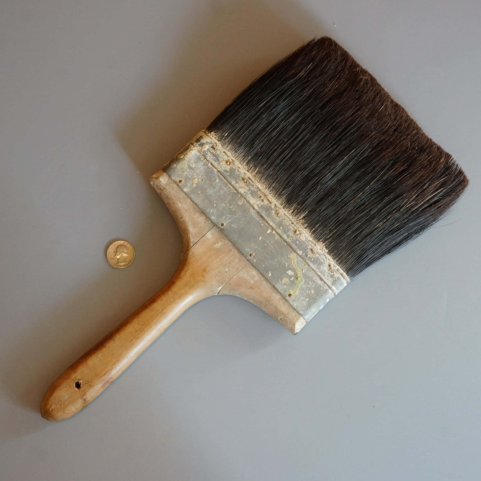 Paint Brush 桌布 壁紙 — 一個用過的畫筆，帶有木柄和金屬箍，放在一個灰色平面上，旁邊有一枚金幣。