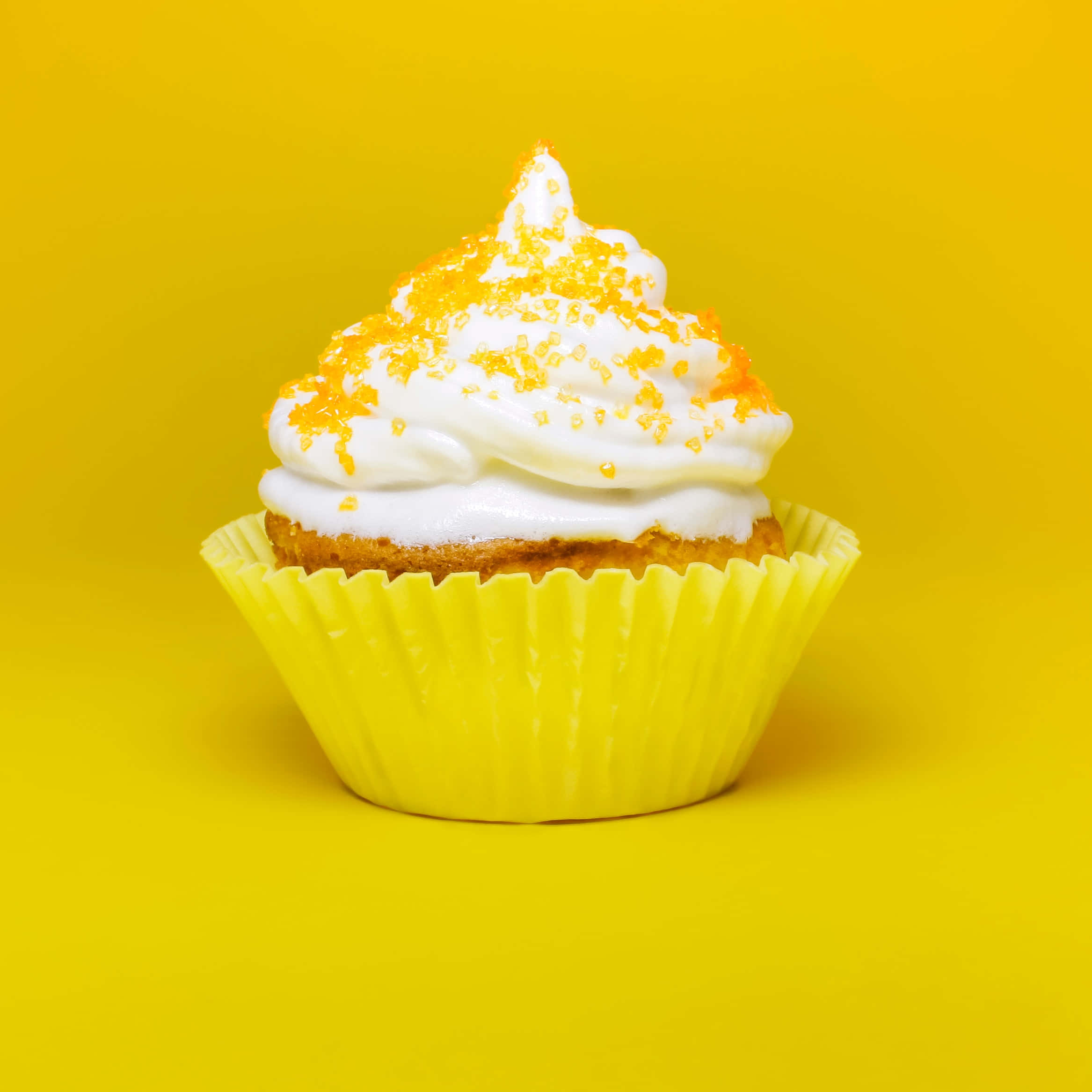 Cupcake 桌布 壁紙 — 一個用黃色紙杯盛裝的杯子蛋糕，上面有白色糖霜和橙色糖粒，背景是純黃色。