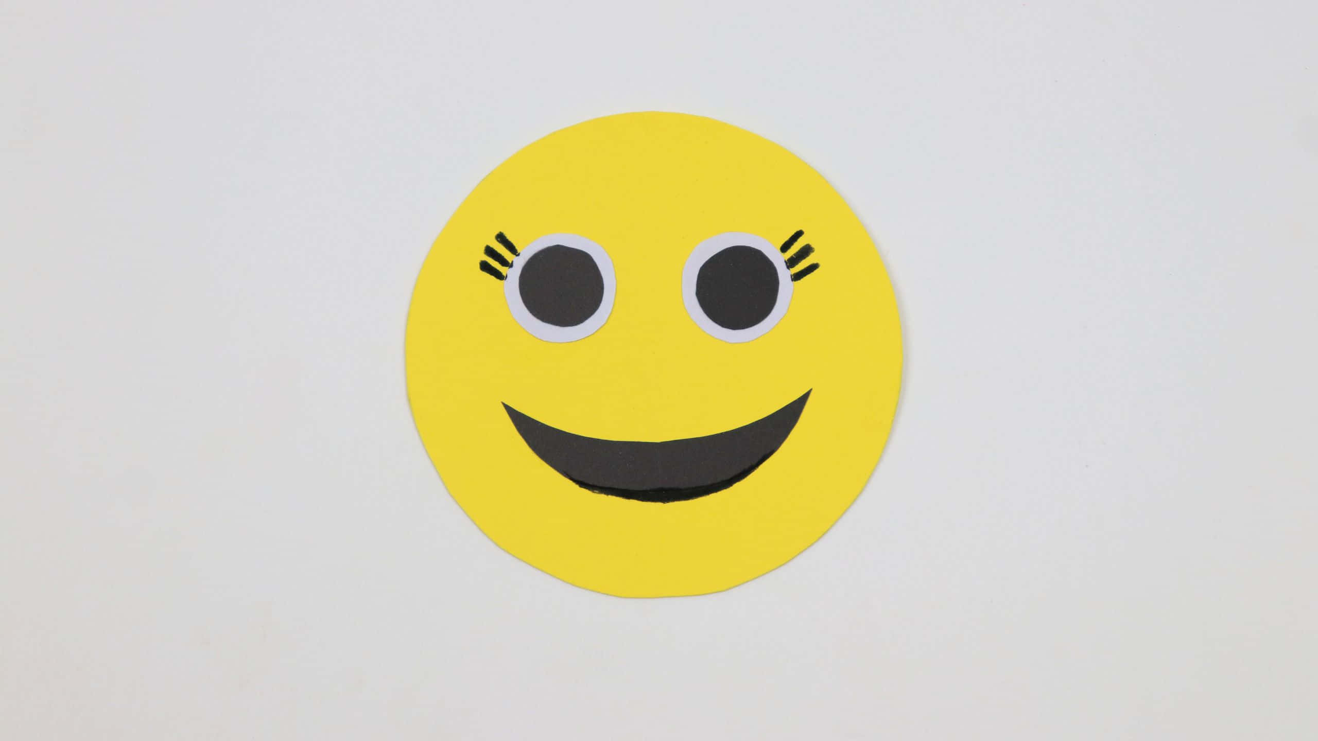 Smile Emoji 桌布 壁紙 — 一個用黃色紙製成、帶有黑色眼睛和睫毛的笑臉，在純白色背景上微笑。