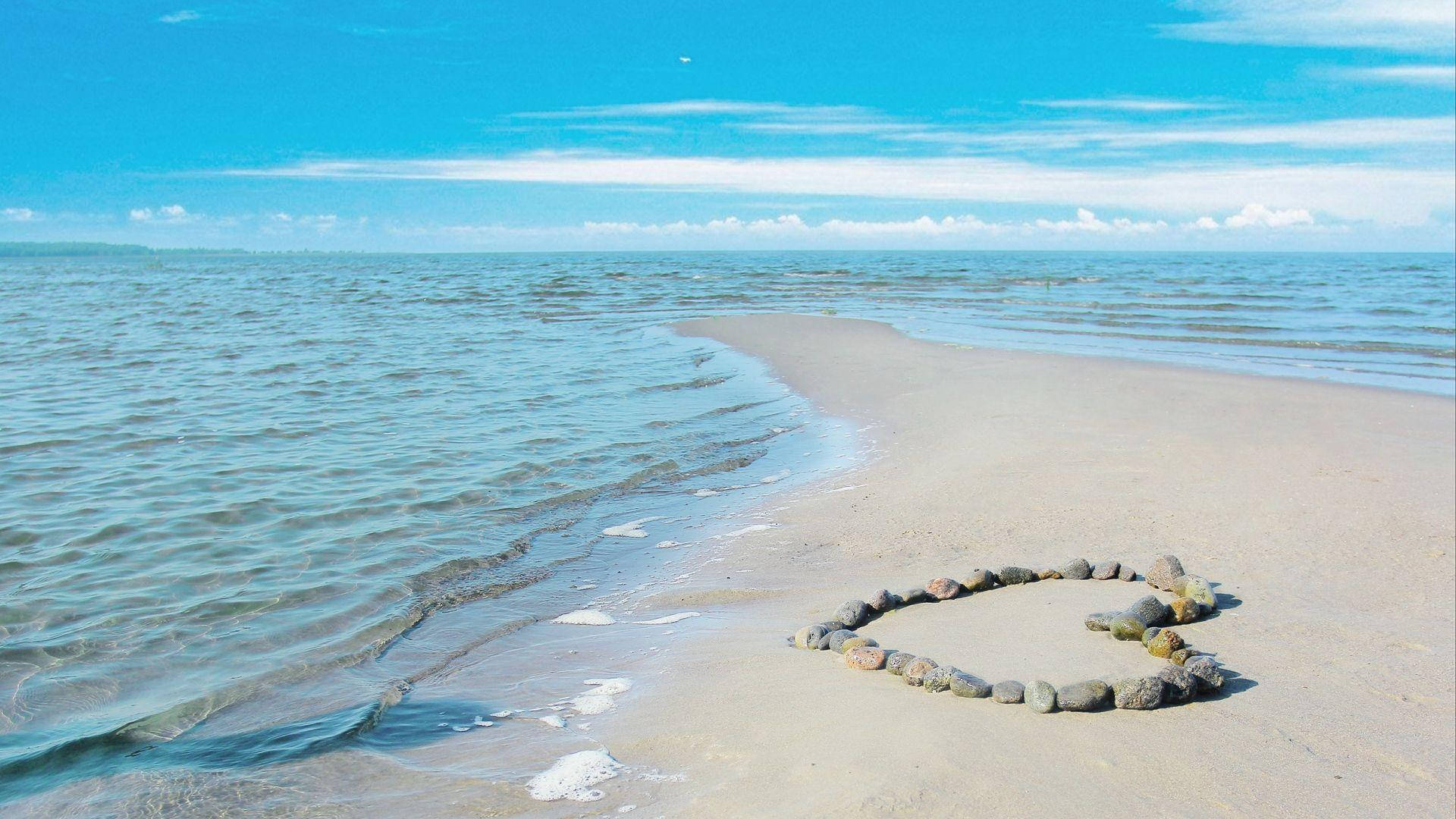 Love Nature 桌布 壁紙 — 一個由光滑石頭組成的愛心形狀，放置在陽光明媚的沙灘上，在清澈的藍天下，溫柔的海浪拍打著海岸。