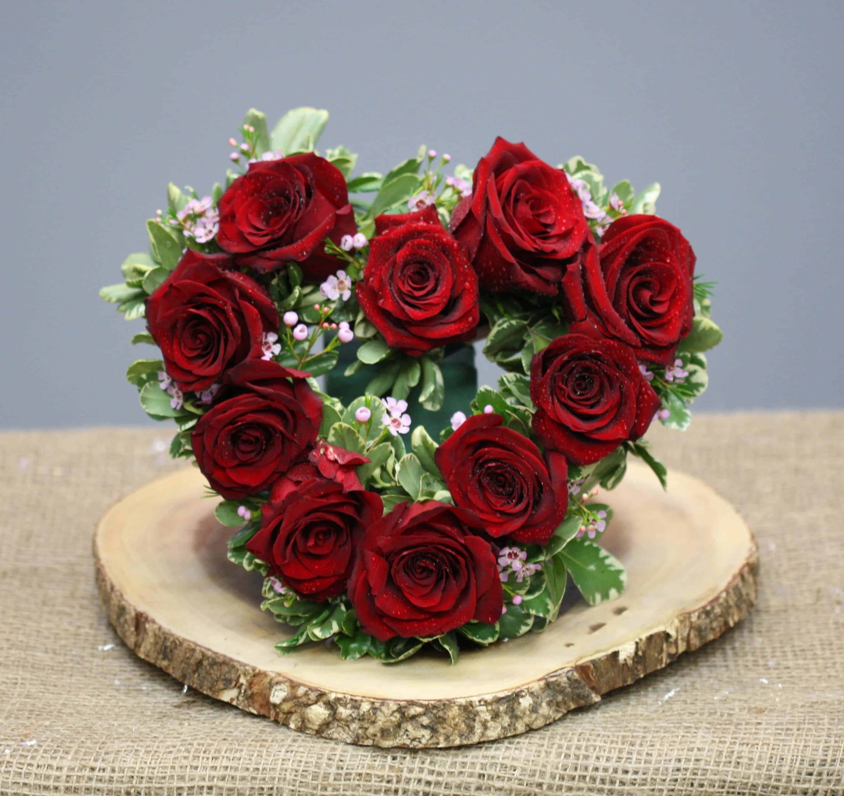 Rose Heart 桌布 壁紙 — 一個由深紅玫瑰、綠葉和小白花組成的心形佈置，放在粗樸木片上，底下是麻布。