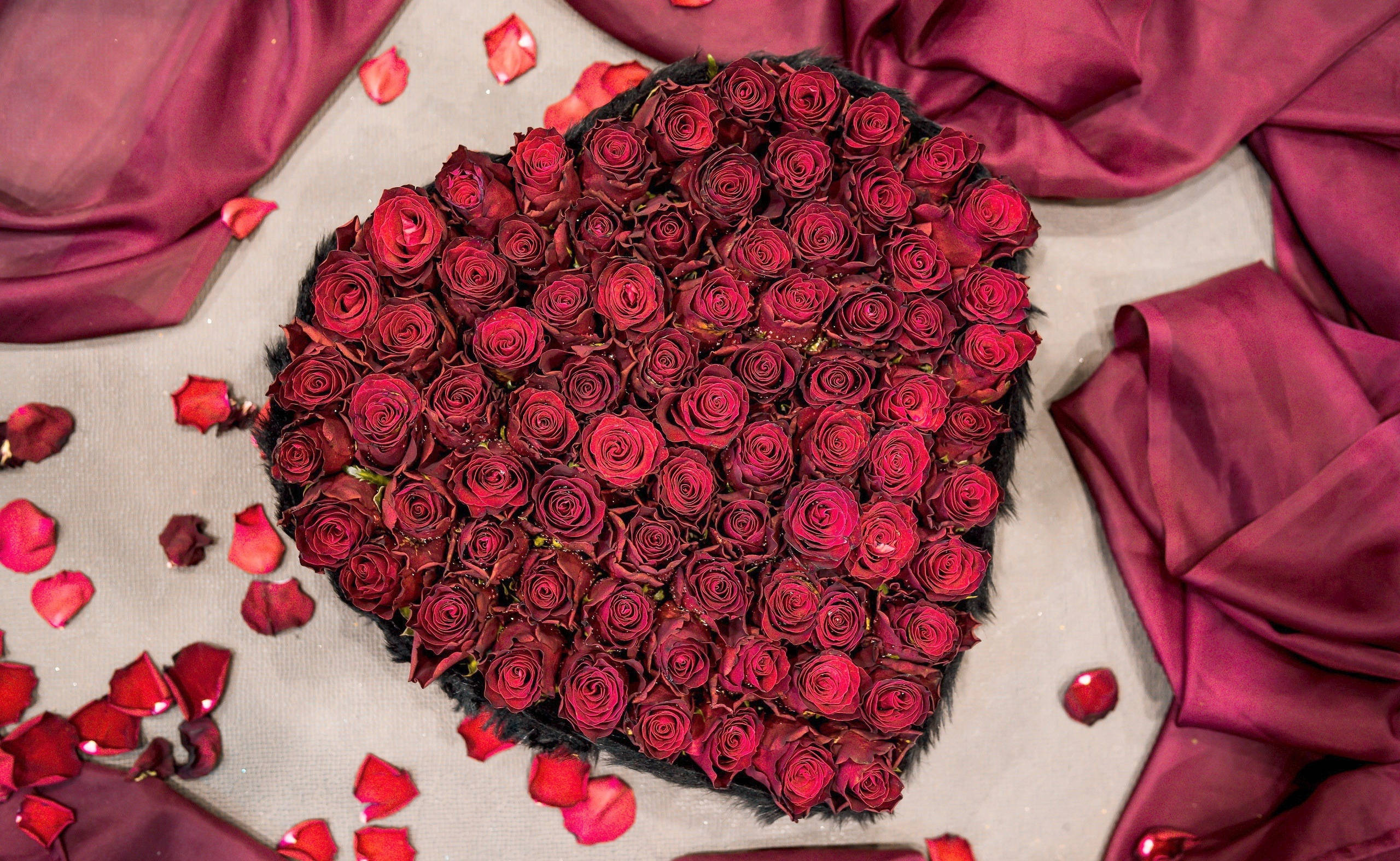 Romantic Rose 桌布 壁紙 — 一個由深紅玫瑰組成的心形佈置，周圍環繞著酒紅色布料和散落的花瓣，置於淺色表面上。