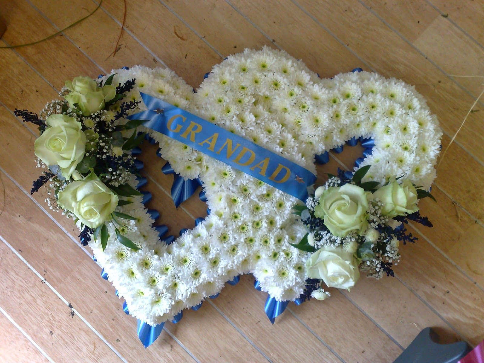 Funeral 桌布 壁紙 — 一個由白色菊花和淺綠色玫瑰製成的心形花卉致敬品放在木地板上，上面有一條寫著GRANDAD的藍色絲帶。