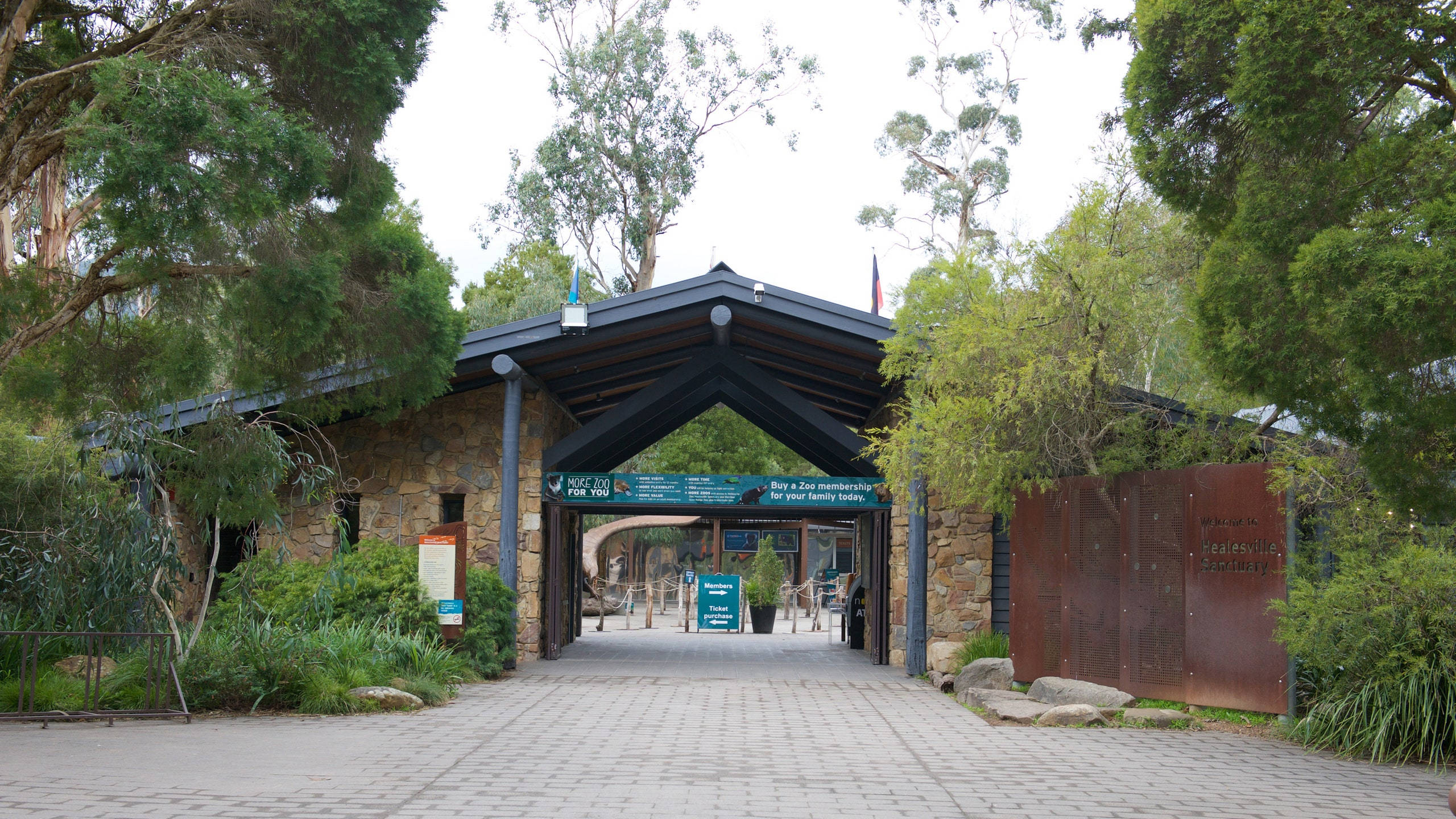 墨爾本 桌布 壁紙 — 一個由石頭和木材建造的動物園入口拱門，被茂密的綠樹和鋪砌的小徑環繞，天空多雲。