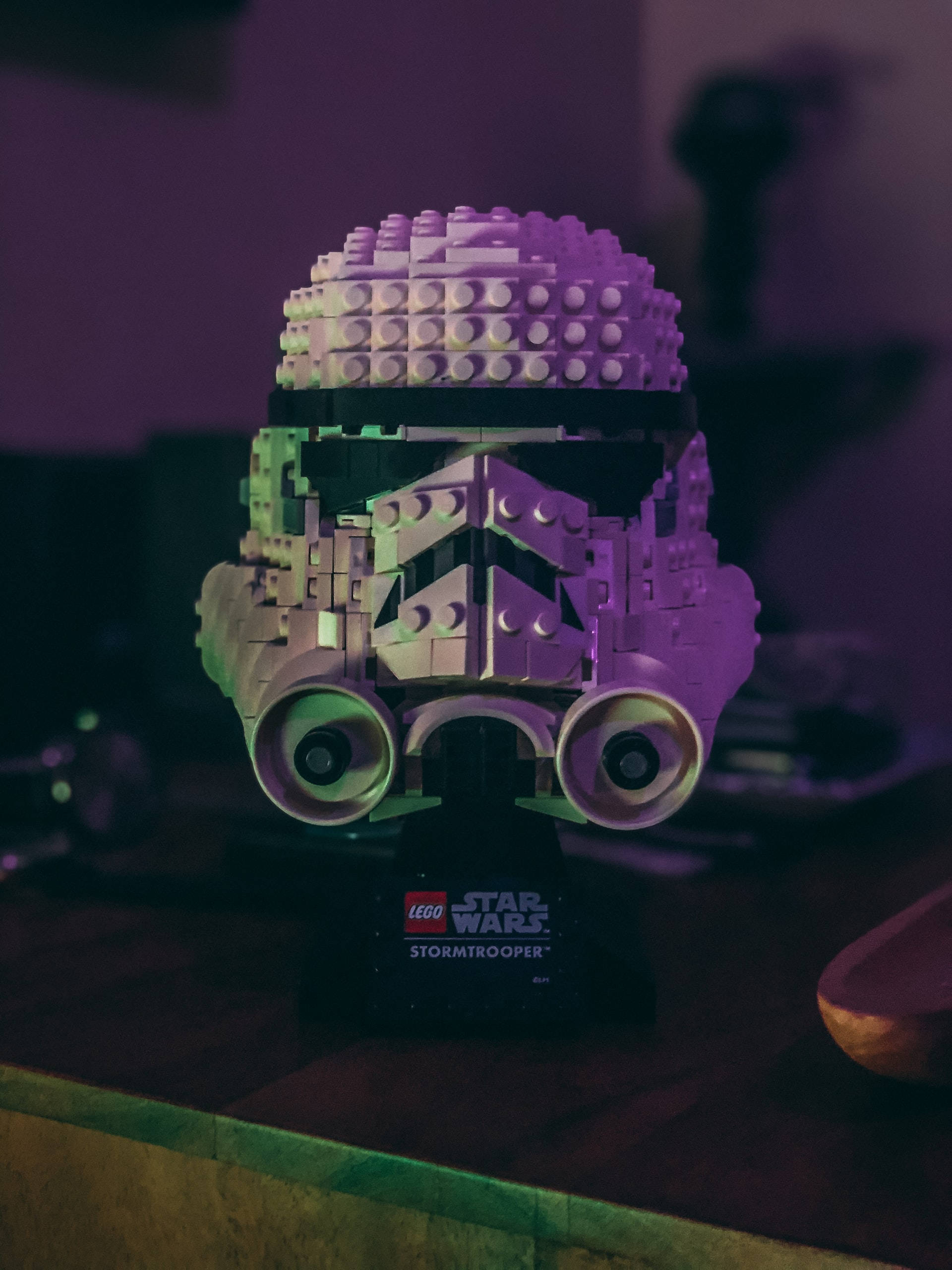 星際大戰 iPhone 桌布 壁紙 — 一個由紫色和綠色燈光照亮的LEGO Star Wars 風暴兵頭盔模型，放置在昏暗房間的展示架上。