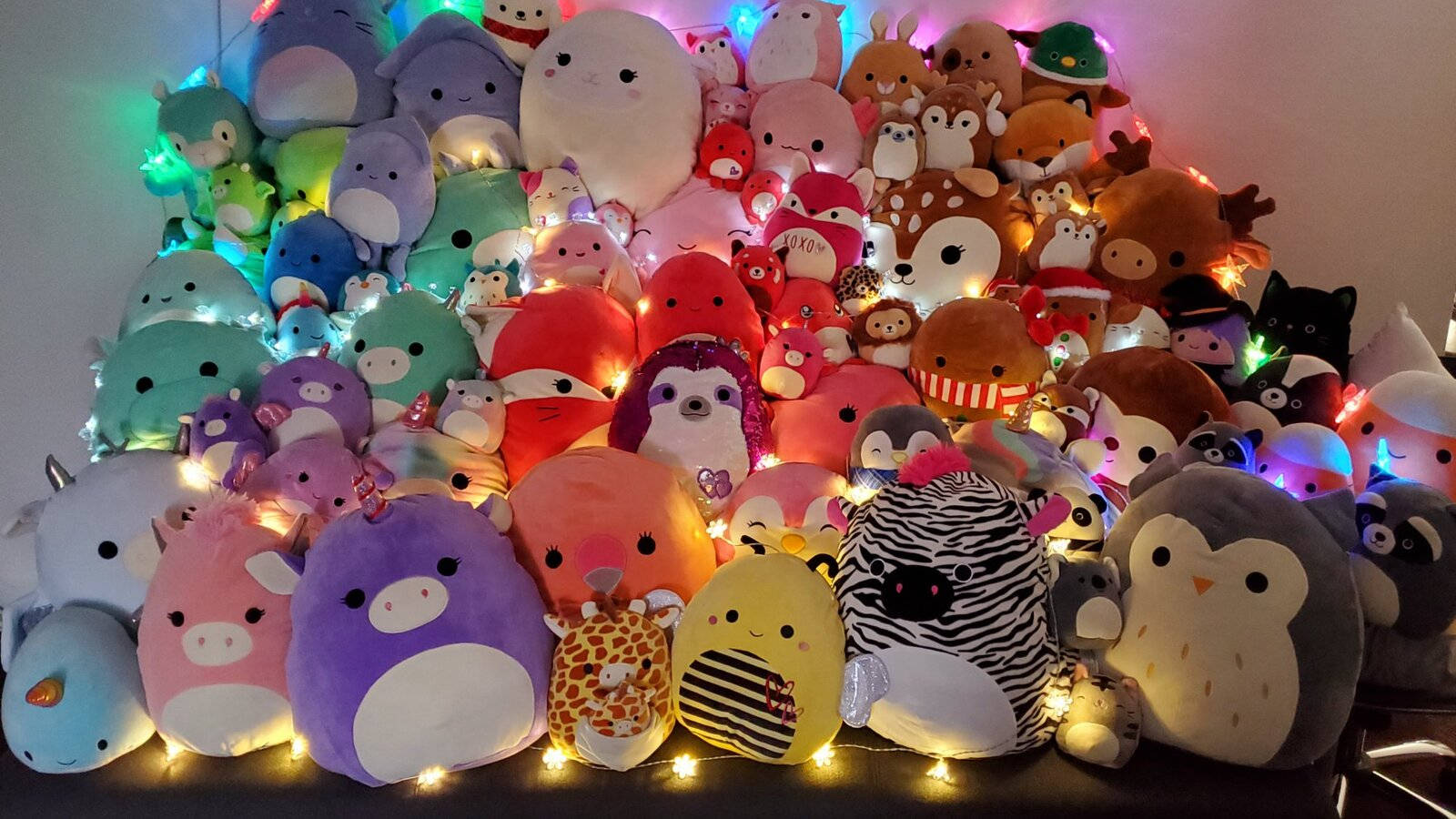 一個由色彩繽紛的毛絨玩具堆成的金字塔，被串燈照亮。 — Squishmallows 壁紙