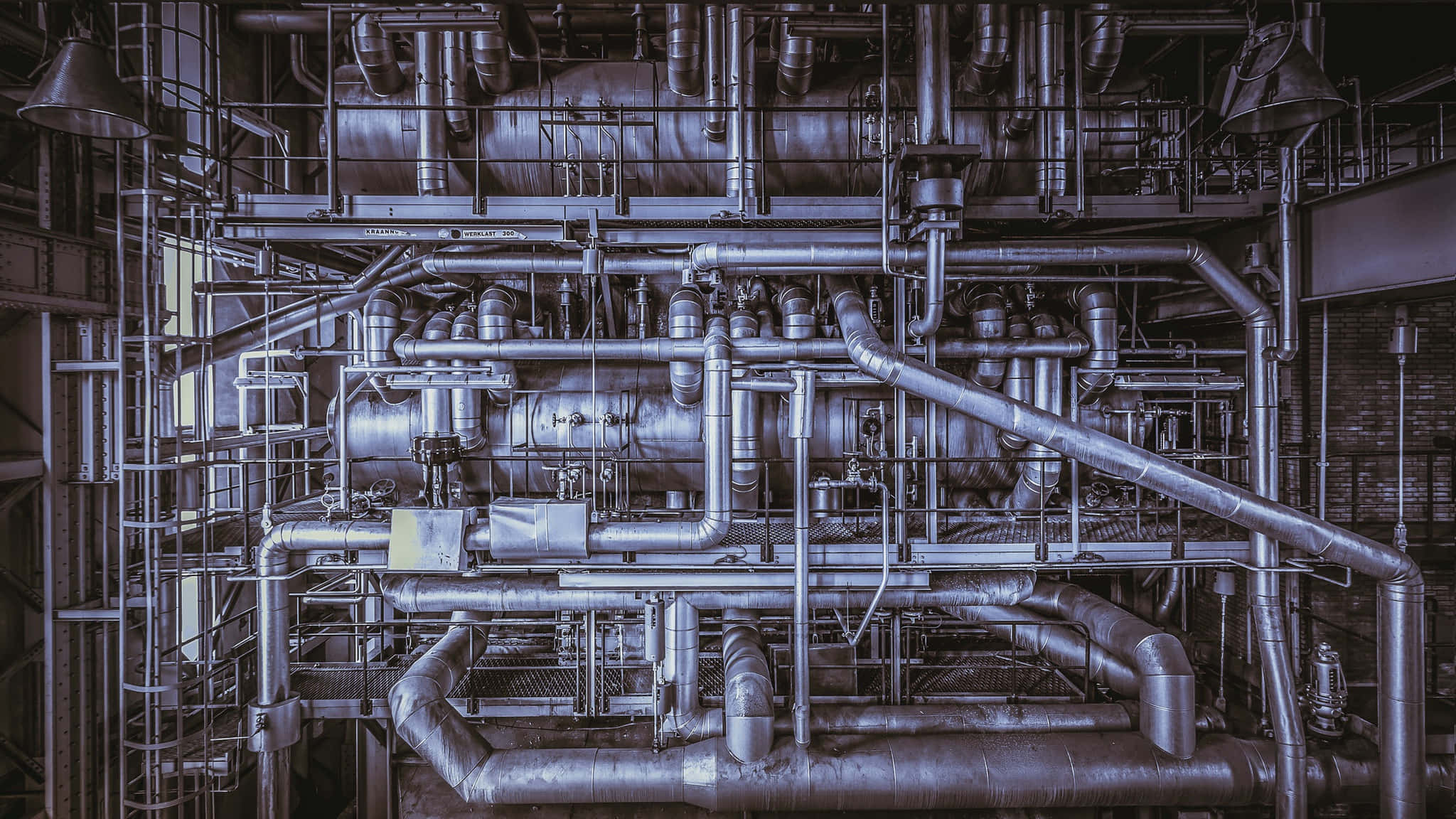 Plumbing 桌布 壁紙 — 一個由金屬管道和工業機械組成的複雜網絡，充滿了昏暗的工廠內部，呈現出一種粗獷的機械美學。
