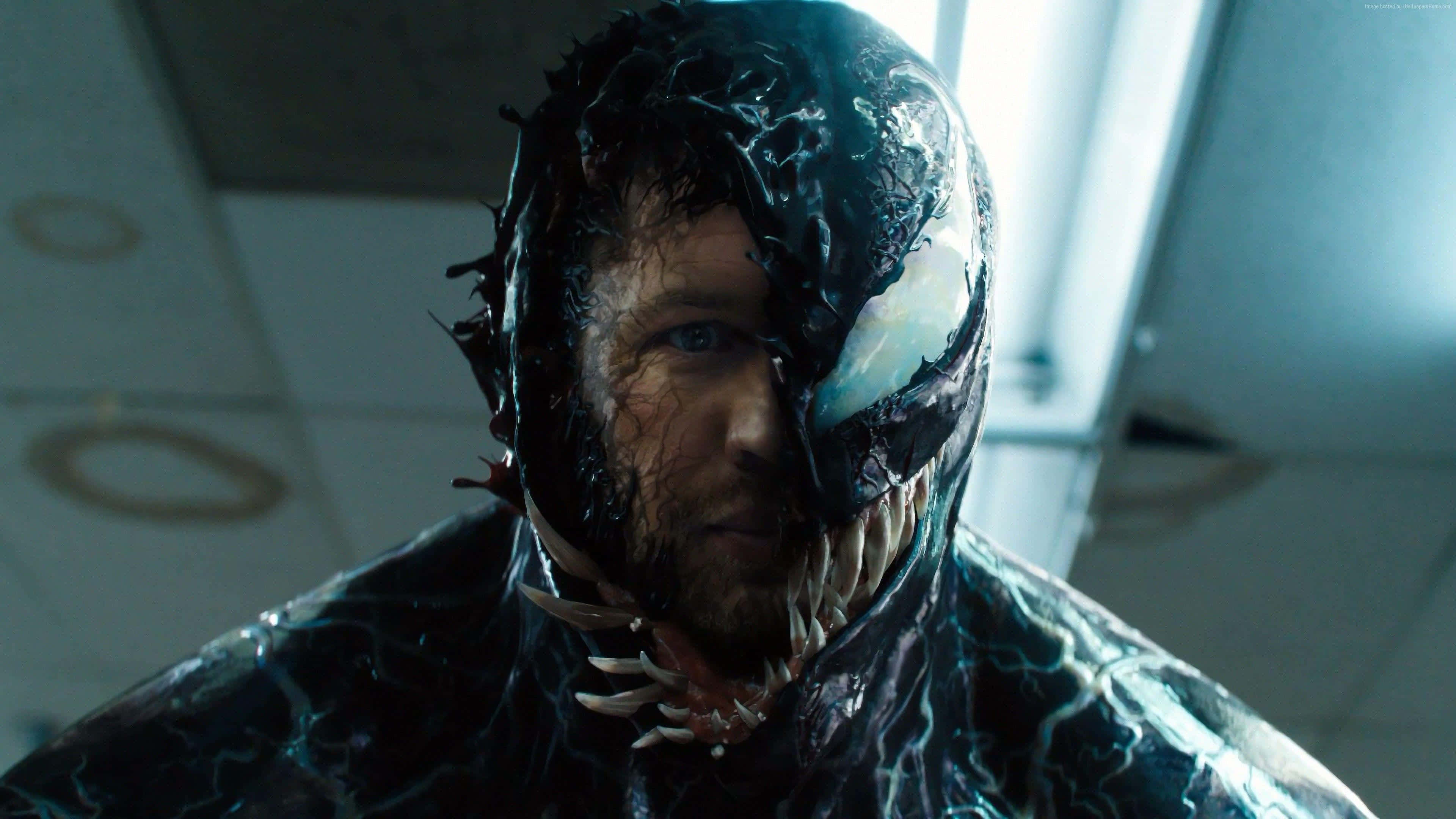 Tom Hardy Venom 桌布 壁紙 — 一個男人半變身成黑色共生體猛毒的特寫，露出尖牙和發光的白色眼睛，身處無菌室。