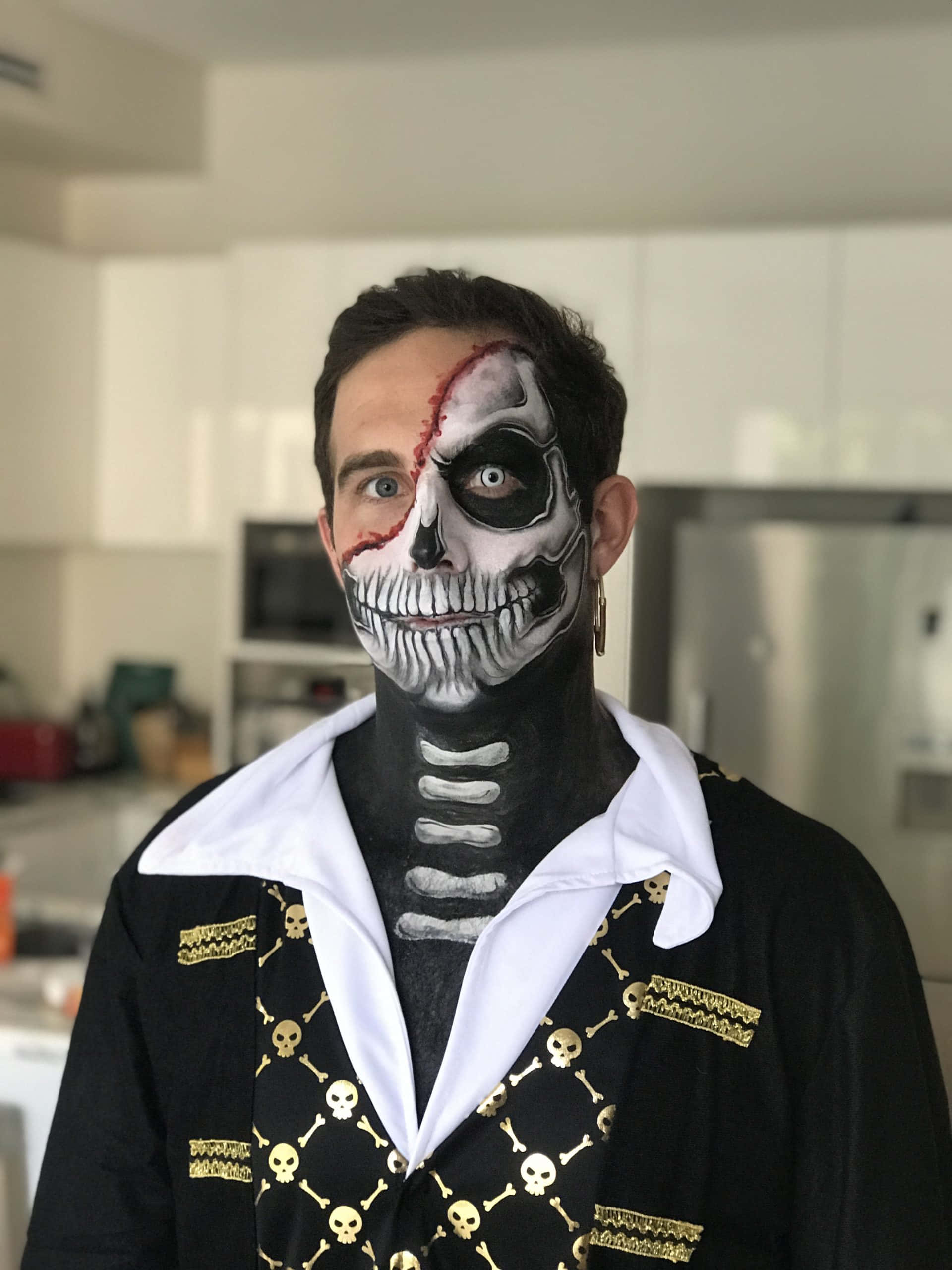 Halloween Face Paint 桌布 壁紙 — 一個男人半邊臉畫成骷髏，另一半自然，穿著骷髏圖案服裝站在廚房裡。