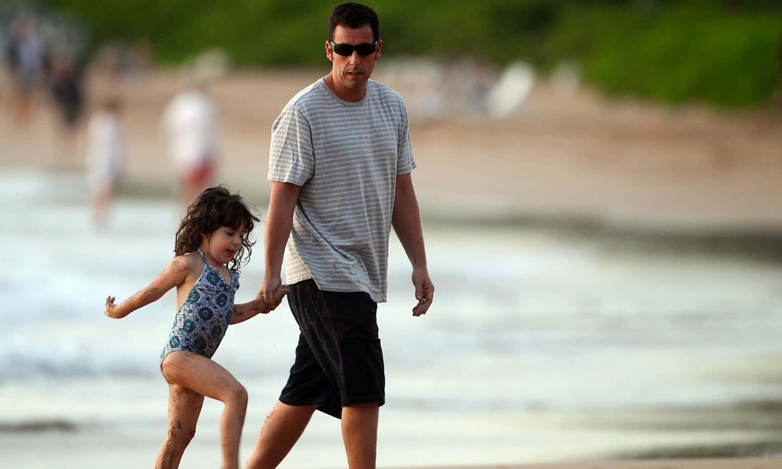 Adam Sandler 桌布 壁紙 — 一個男人和一個年輕女孩手牽手沿著陽光明媚的海灘散步，溫柔的海浪拍打著他們的腳。