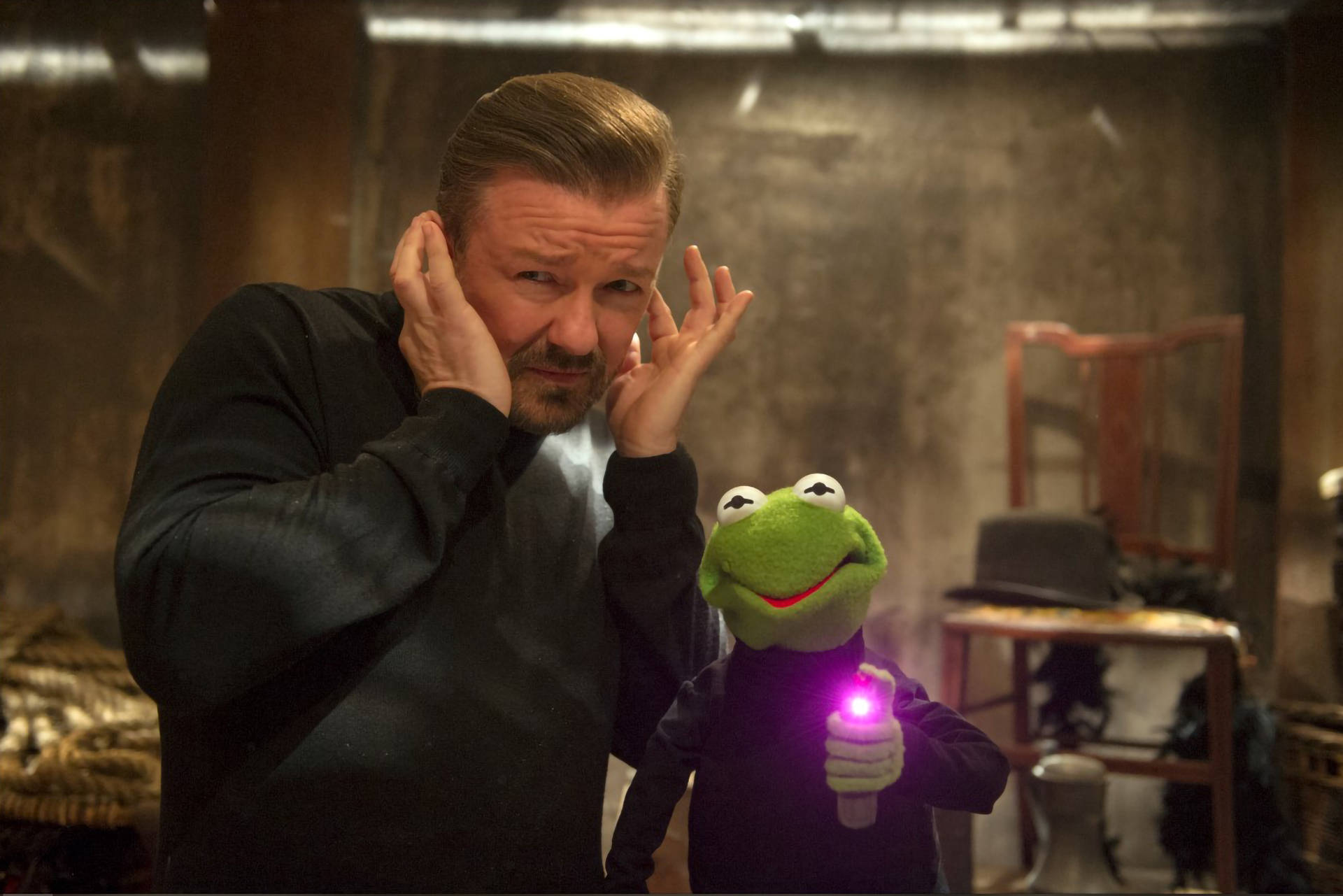 muppets 桌布 壁紙 — 一個男人和青蛙柯密特都捂住耳朵，柯密特在昏暗的倉庫裡拿著一個發光的紫色手電筒。