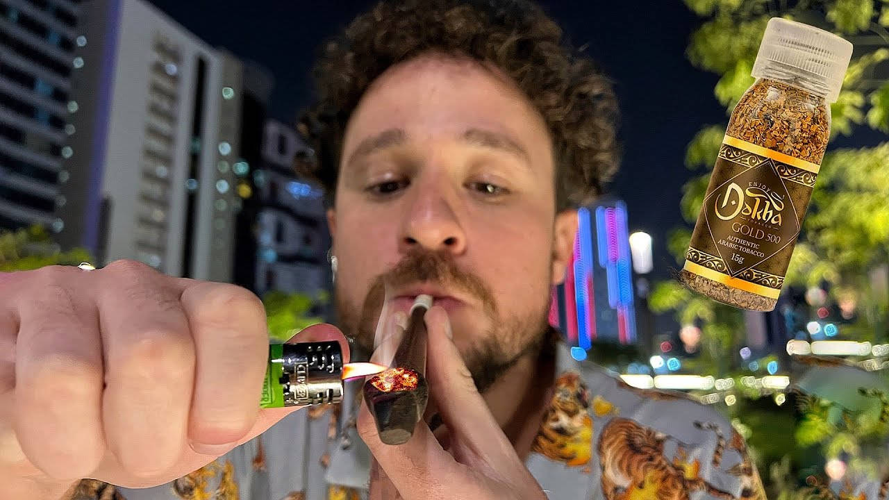 Qatar 桌布 壁紙 — 一個男人在充滿活力的夜間城市景觀中點燃一支大麻煙，背景有發光的霓虹燈和一個漂浮的Dekka Gold 500瓶子。