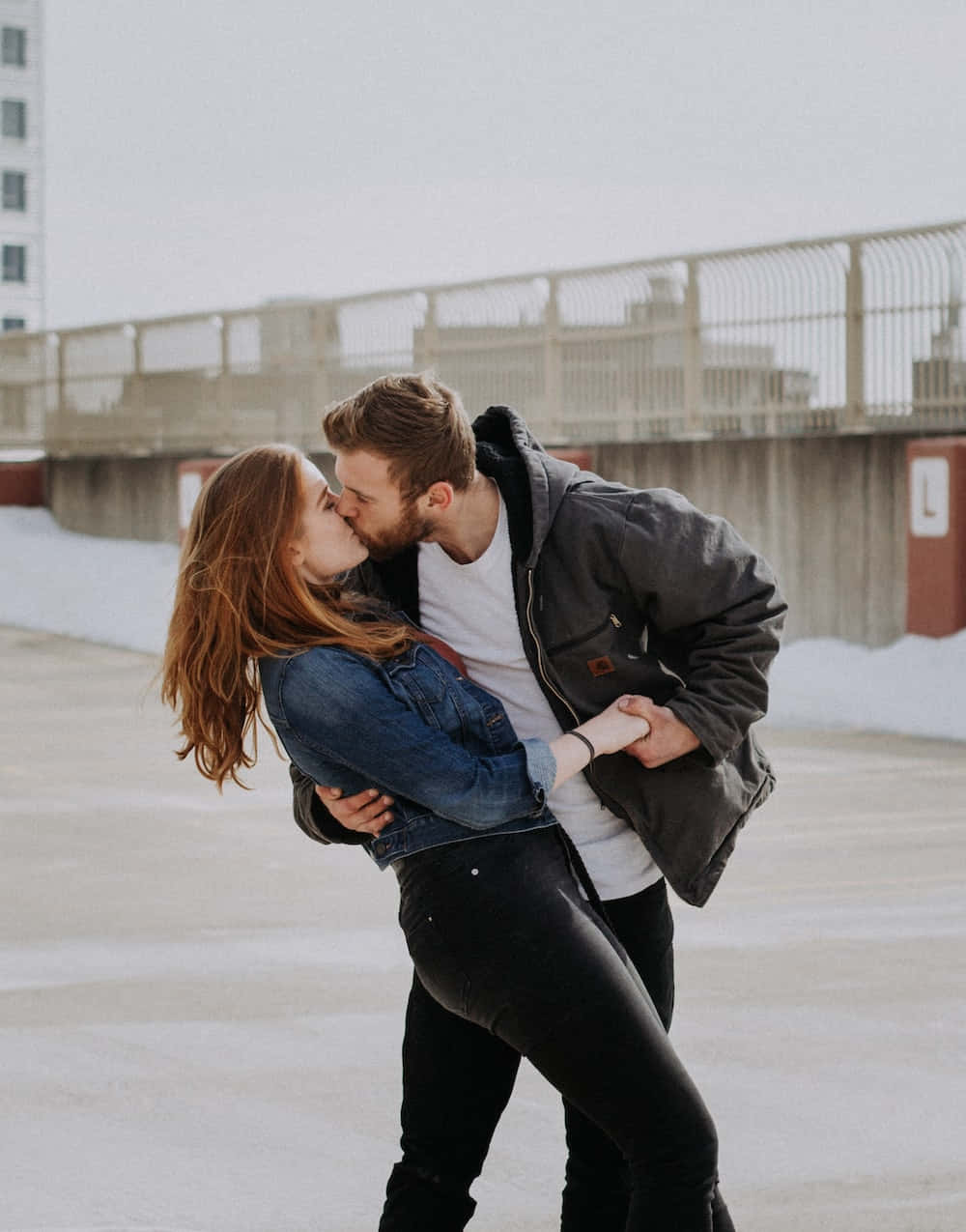Kissing 桌布 壁紙 — 一個男人在冬日擁抱中傾斜一個女人，他們在雪景城市屋頂上熱情接吻，背景有建築物。
