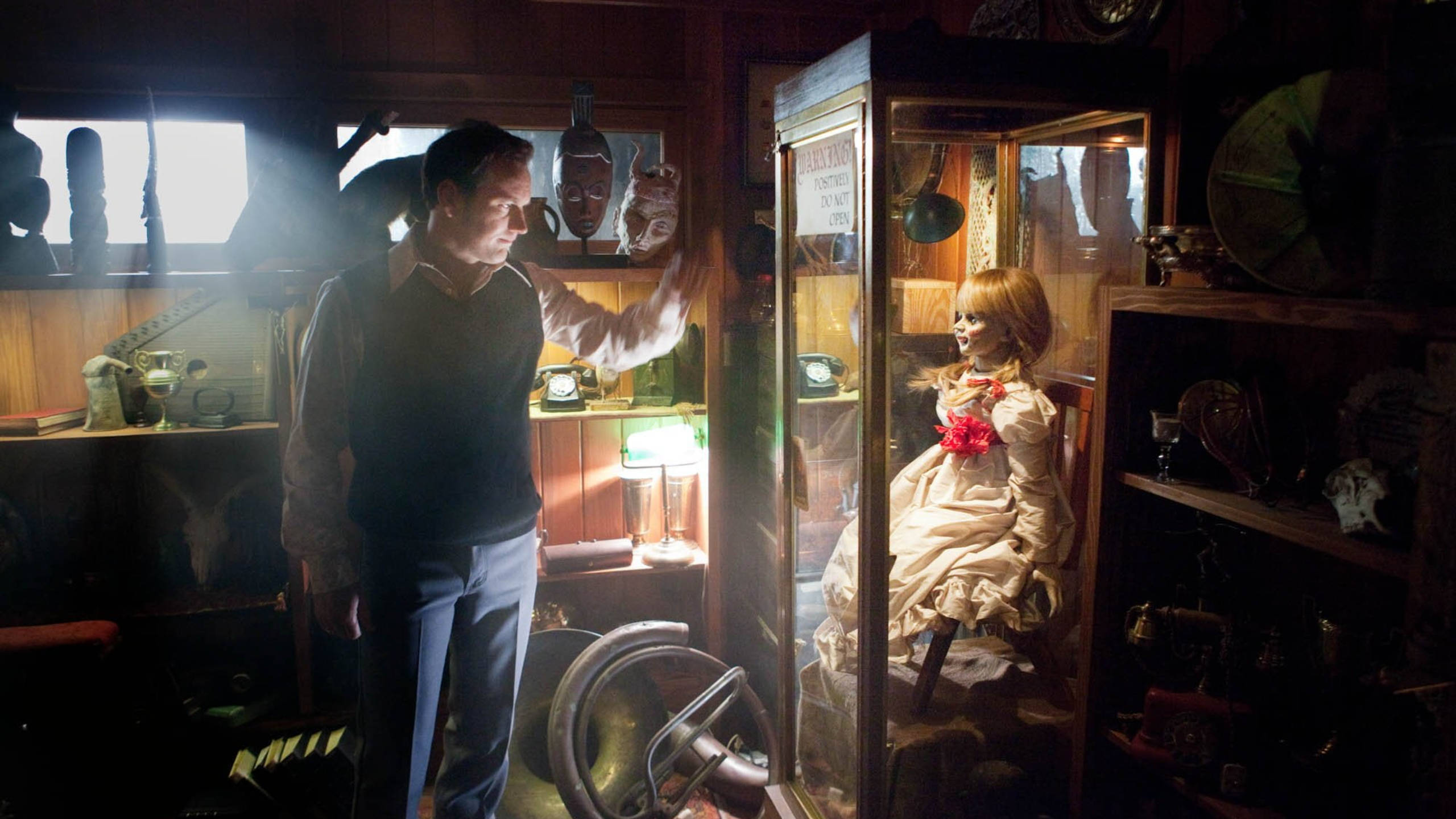 Annabelle 桌布 壁紙 — 一個男人在昏暗雜亂的房間裡伸手朝向一個玻璃櫃，裡面有一個帶紅花的恐怖娃娃。