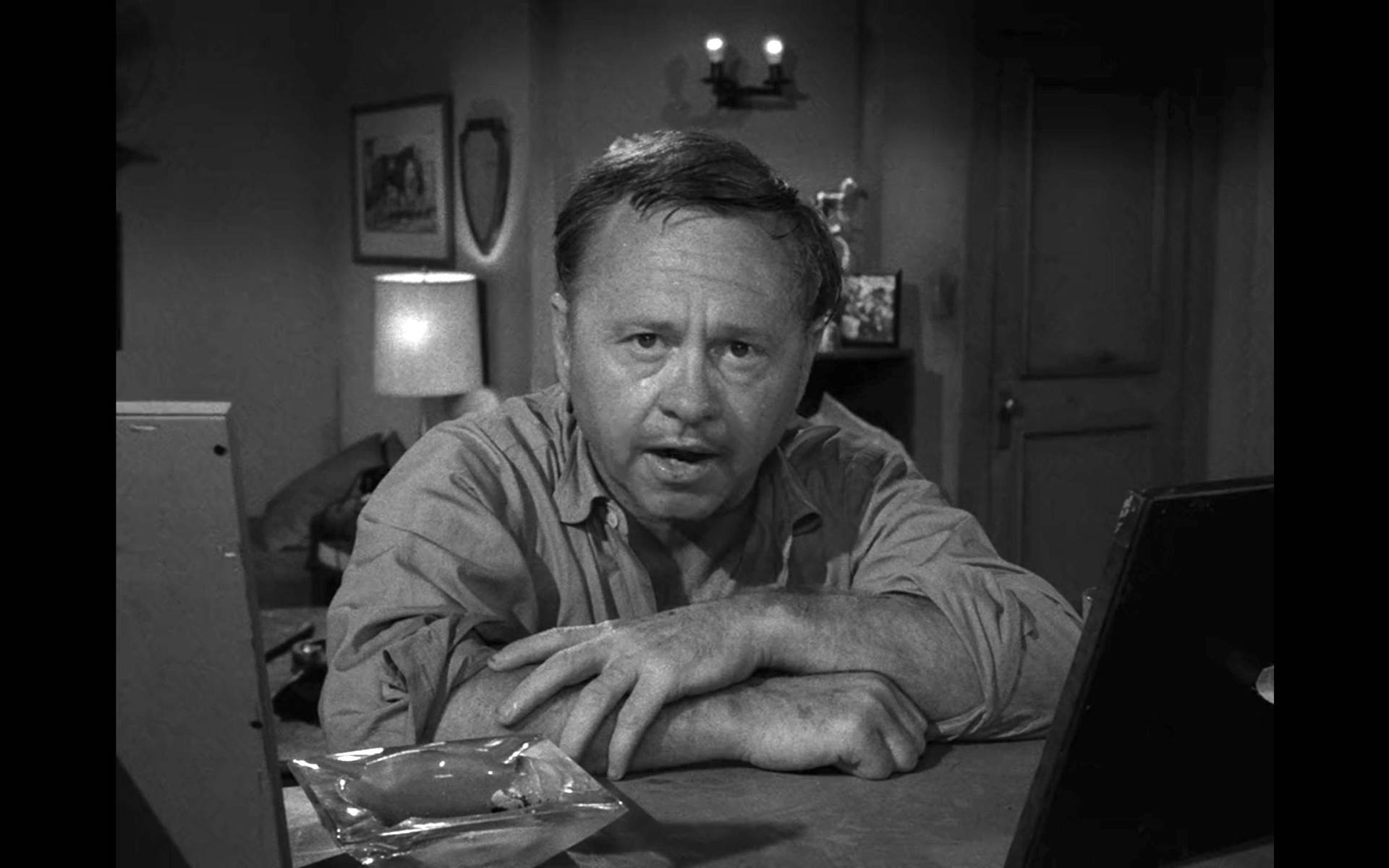 mickey rooney 桌布 壁紙 — 一個男人在燈光昏暗的房間裡，桌子上有燈和牆上掛著裱框的圖片，身體前傾。