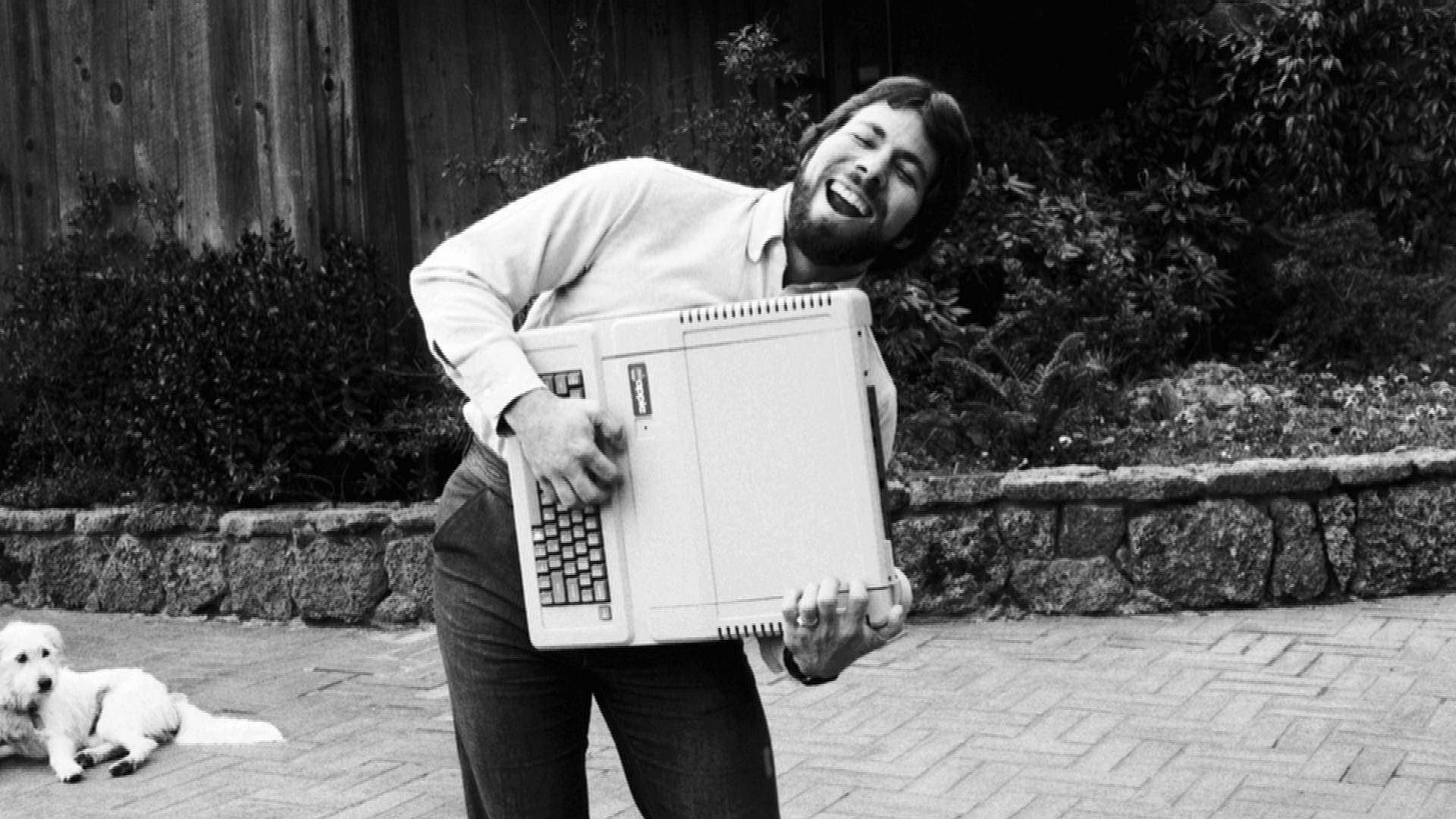 黑白 桌布 壁紙 — 一個男人在磚砌露台戶外開心笑著，拿著一部復古Apple II電腦，附近有一隻狗。