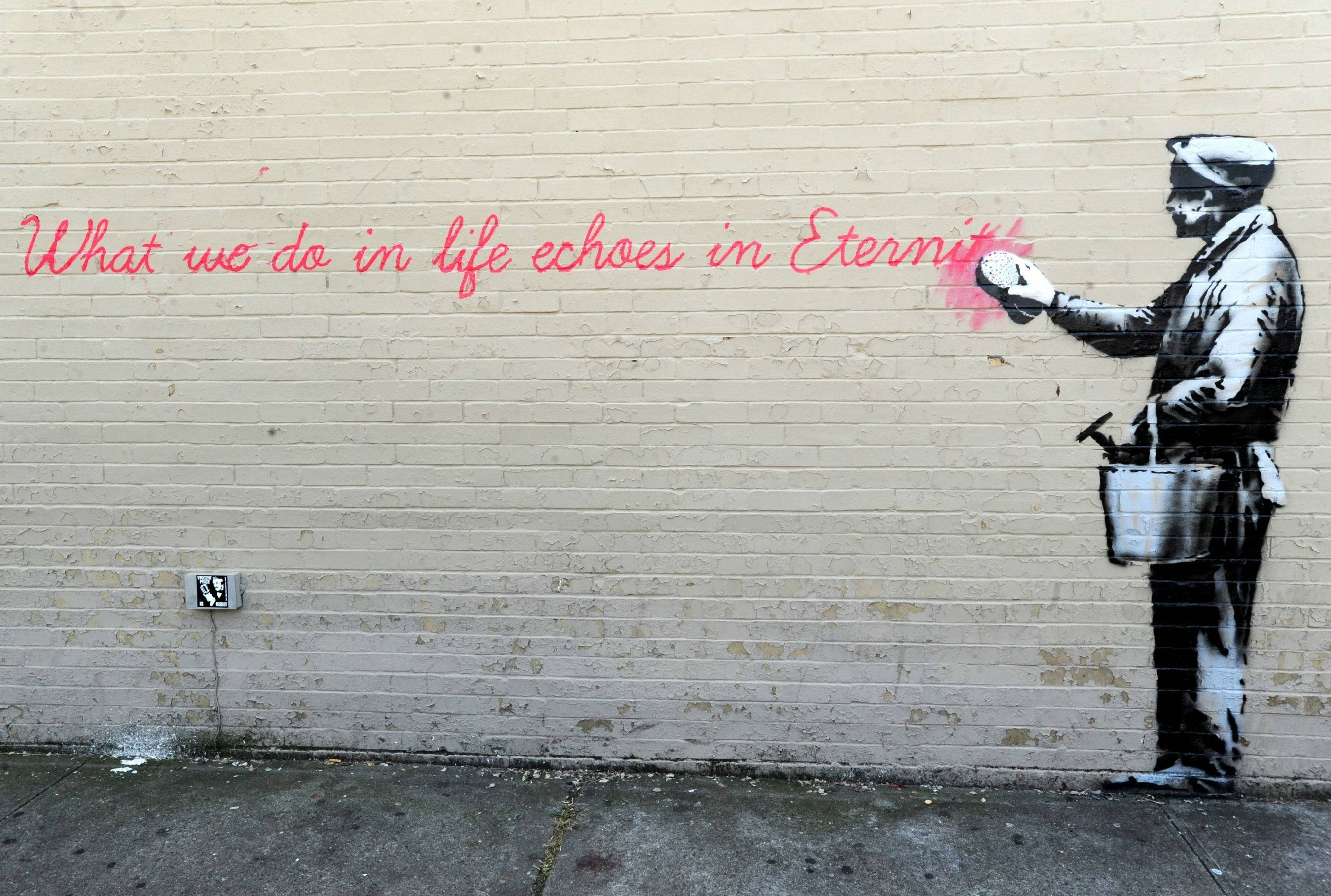 Banksy 桌布 壁紙 — 一個男人在米色磚牆上用粉紅色噴漆「我們在生命中所做的事會在永恆中迴響」的模板塗鴉。