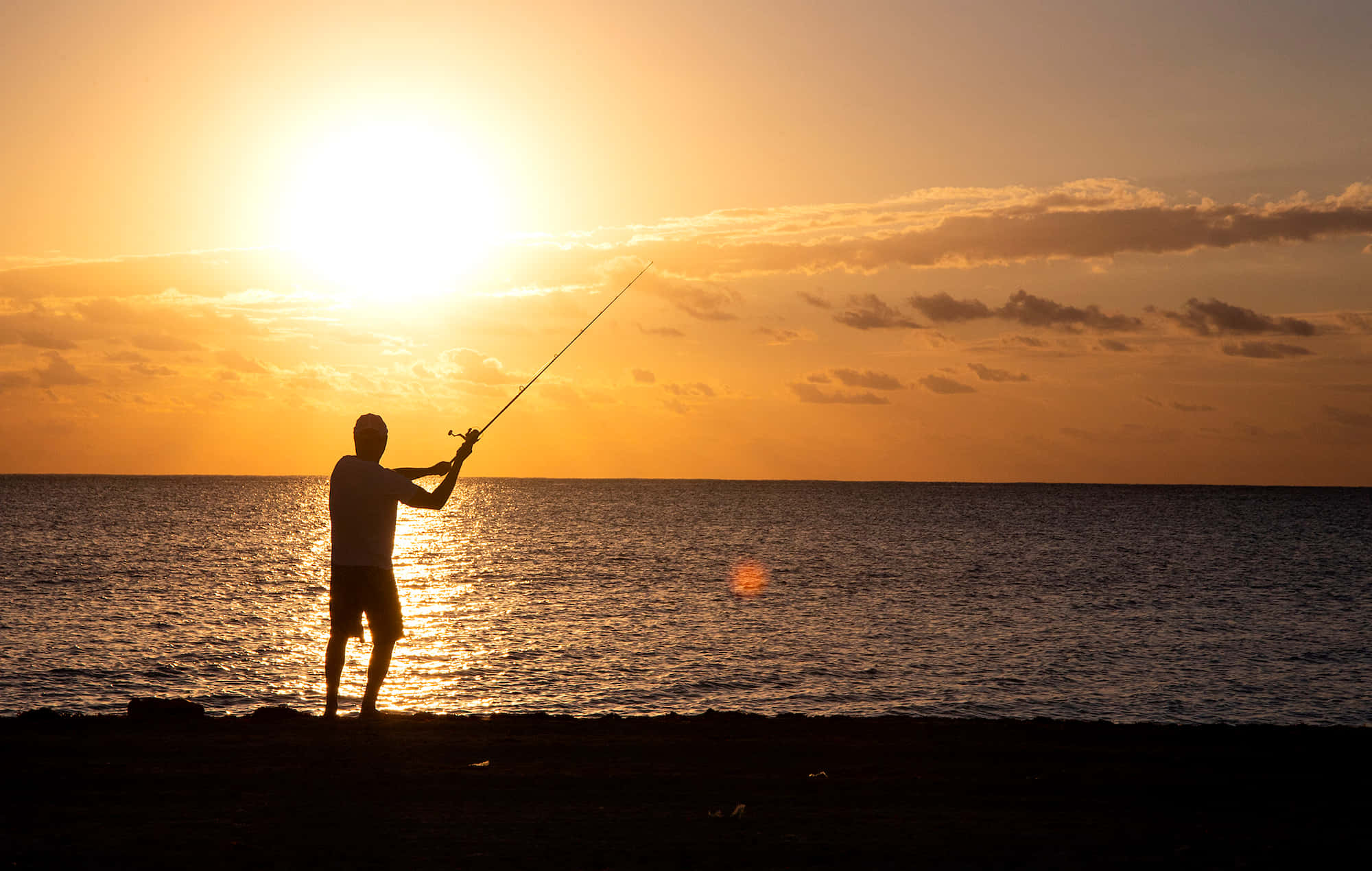 Fishing Rod 桌布 壁紙 — 一個男人在金色日落、雲彩和閃爍海水中拋出釣竿的剪影。