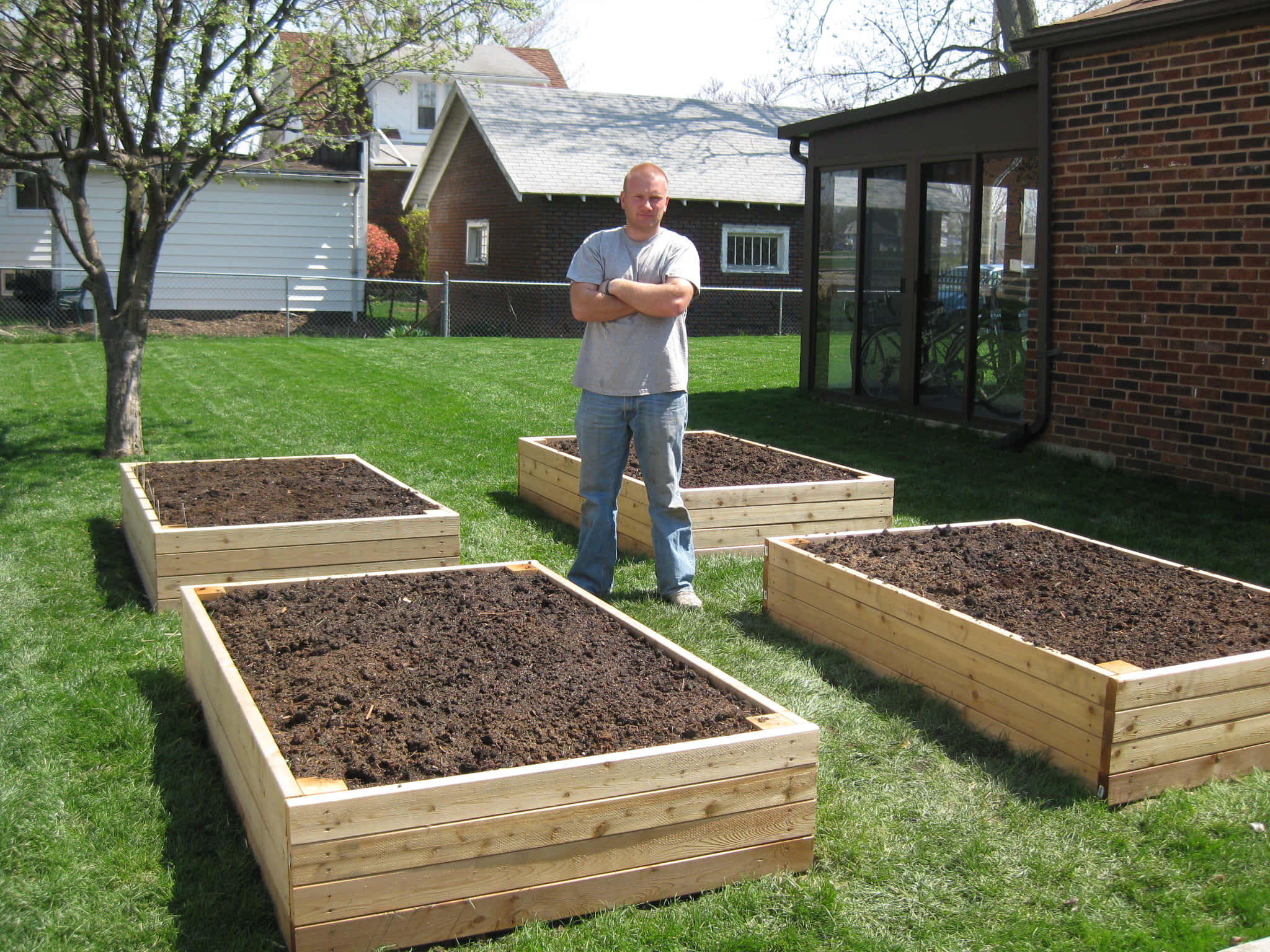 Garden Bed 桌布 壁紙 — 一個男人在陽光明媚的後院雙臂交叉站立，周圍是四個裝滿肥沃土壤的新木製高架花園床。