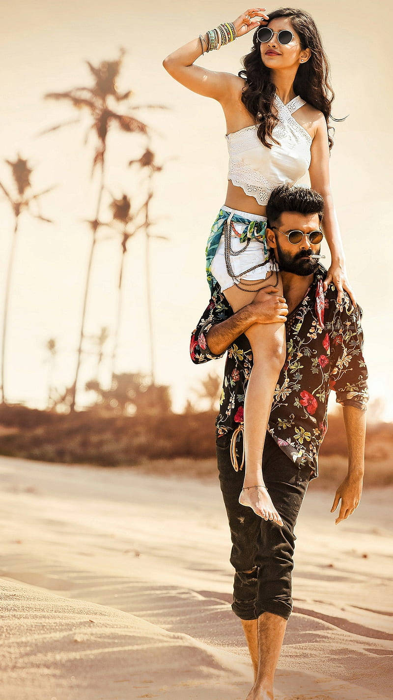 Ram Pothineni 桌布 壁紙 — 一個男人在陽光明媚的海灘上將一個女人扛在肩上，背景有棕櫚樹。