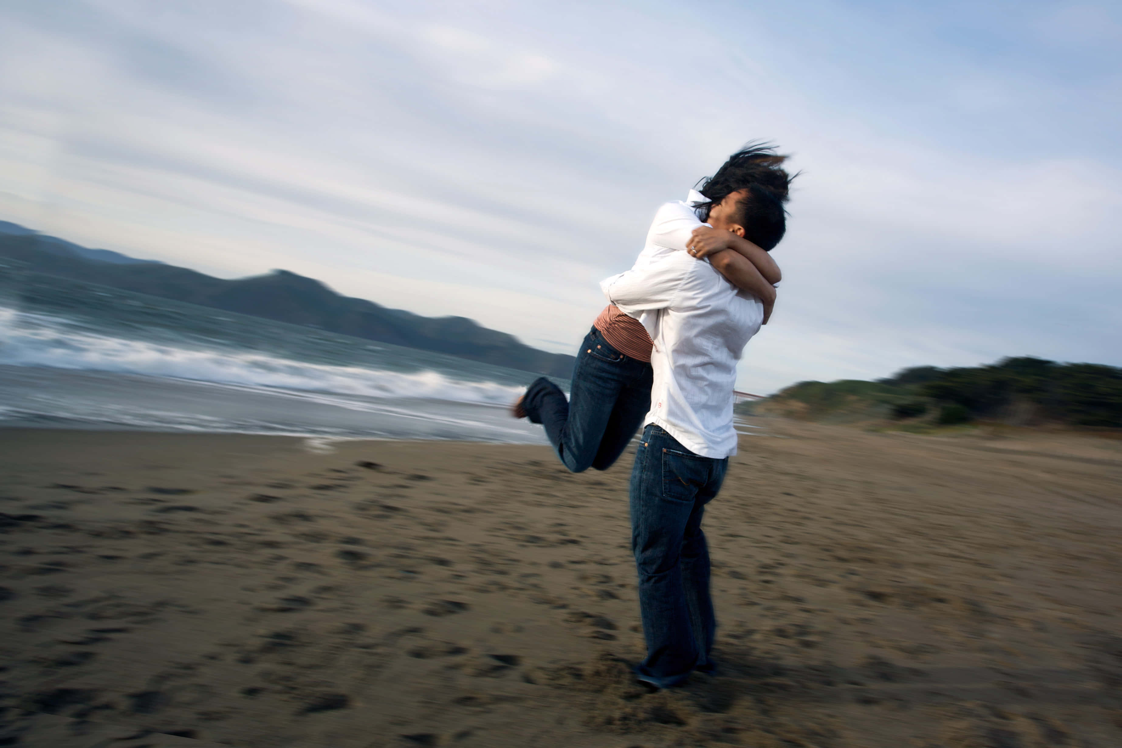 Hug 桌布 壁紙 — 一個男人在風吹沙灘上，快樂地以動態擁抱舉起一個女人，海浪拍打，遠山可見。