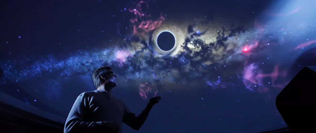 Planetarium 桌布 壁紙 — 一個男人在黑暗的房間裡抬頭凝視著一個令人驚嘆的宇宙投影，其中包含一個黑洞和充滿活力的星雲。