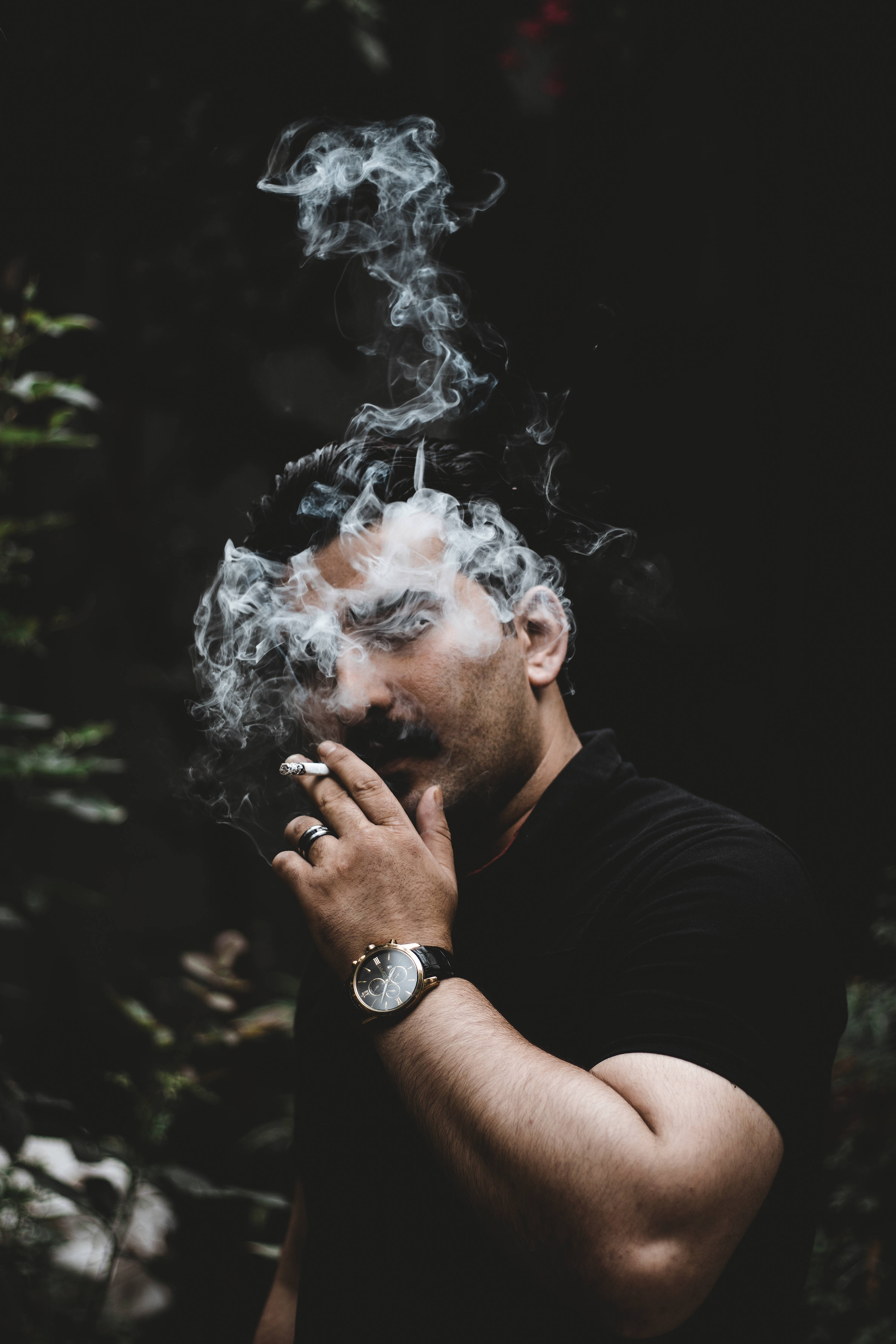 抽菸 桌布 壁紙 — 一個男人在黑暗的森林中呼出濃煙，煙霧在他臉龐周圍旋轉，他手持一支香煙。