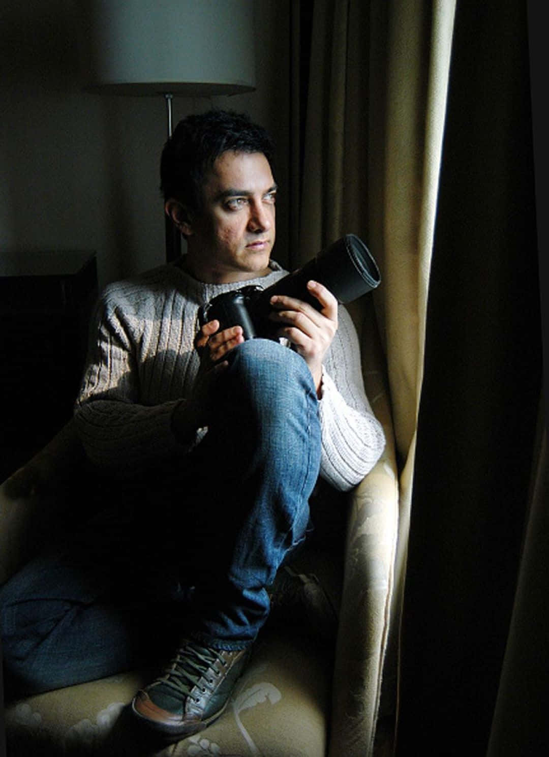 Aamir Khan 桌布 壁紙 — 一個男人坐在光線昏暗的房間裡，手持相機，陽光穿過他身後的窗簾照射進來。
