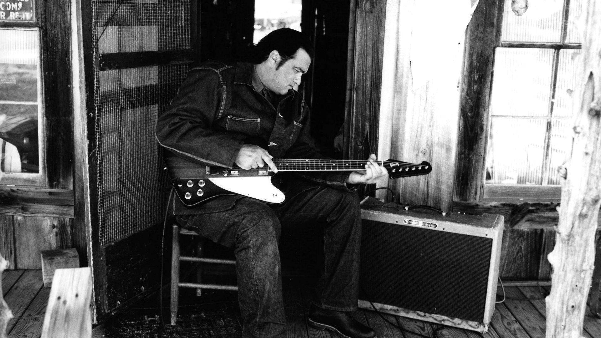 Steven Seagal 桌布 壁紙 — 一個男人坐在凳子上，在質樸的木門廊裡，旁邊有擴音器，彈奏白色電吉他。