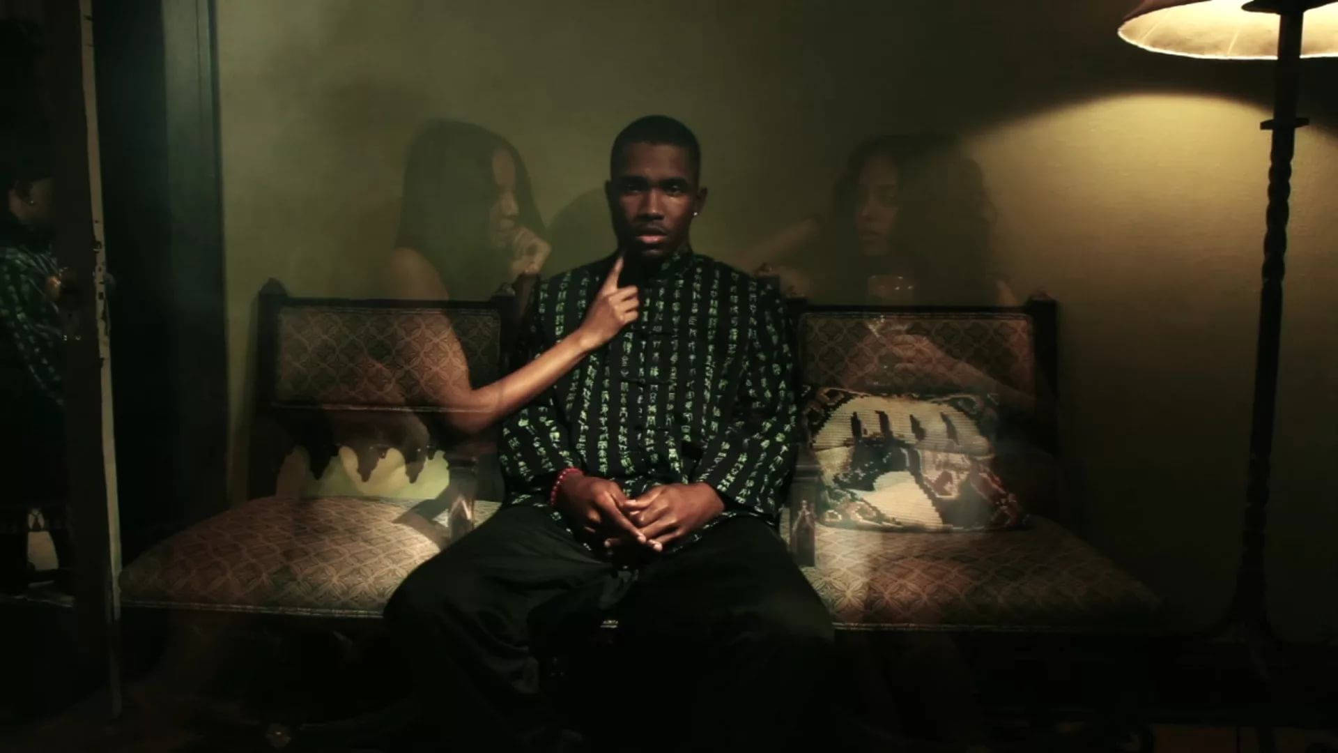 Frank Ocean 桌布 壁紙 — 一個男人坐在圖案沙發上，房間燈光昏暗，身後有幽靈般的女性身影飄浮。